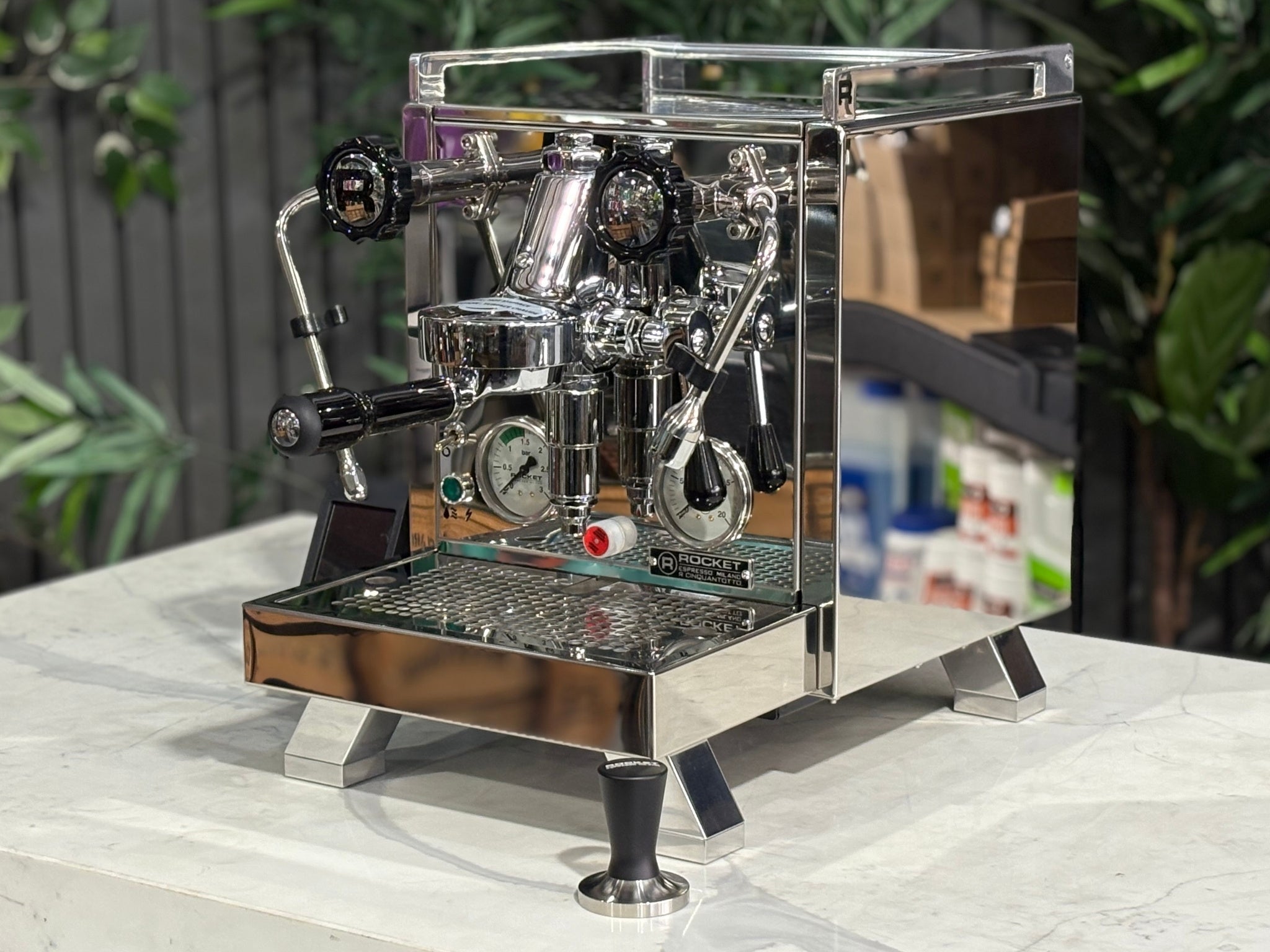Rocket Cinquantotto R58 1 Group Coffee Machine Stainless - New angled view showing the polished stainless steel body, E61 group head, cool touch steam wand, and wide drip tray on a marble bench with greenery in the background.