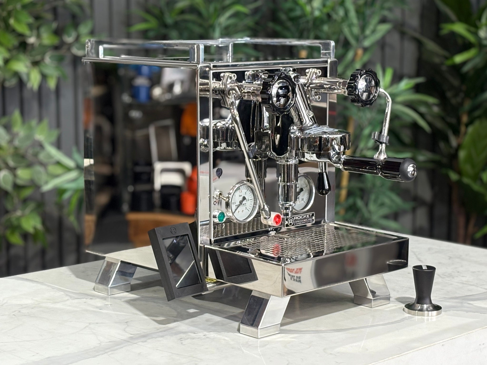 Rocket Cinquantotto R58 1 Group Coffee Machine Stainless - New angled view showing the polished stainless steel body, E61 group head, cool touch steam wand, and wide drip tray on a marble bench with greenery in the background.