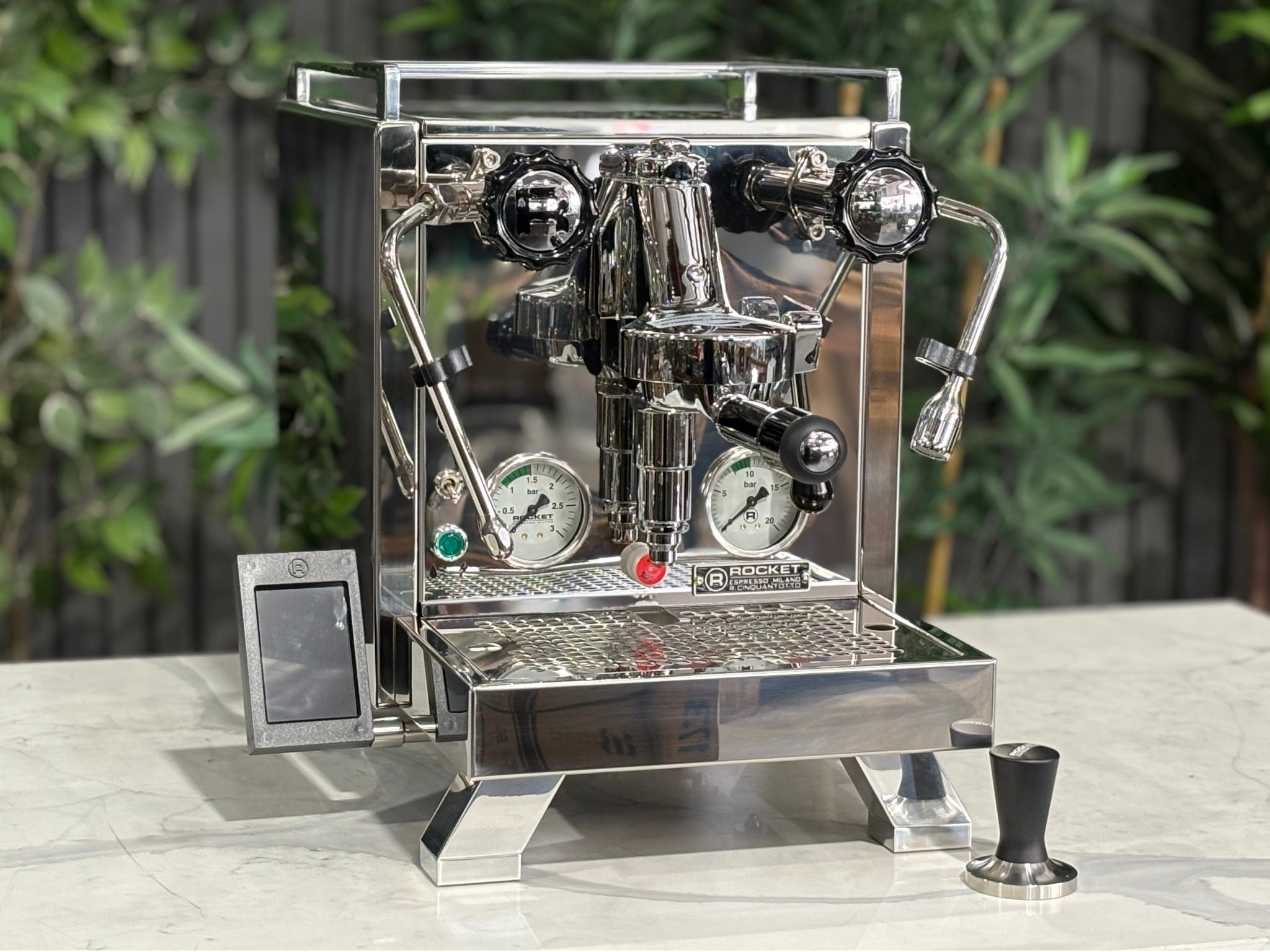 Rocket Cinquantotto R58 1 Group Coffee Machine Stainless - New angled view showing the polished stainless steel body, E61 group head, cool touch steam wand, and wide drip tray on a marble bench with greenery in the background.