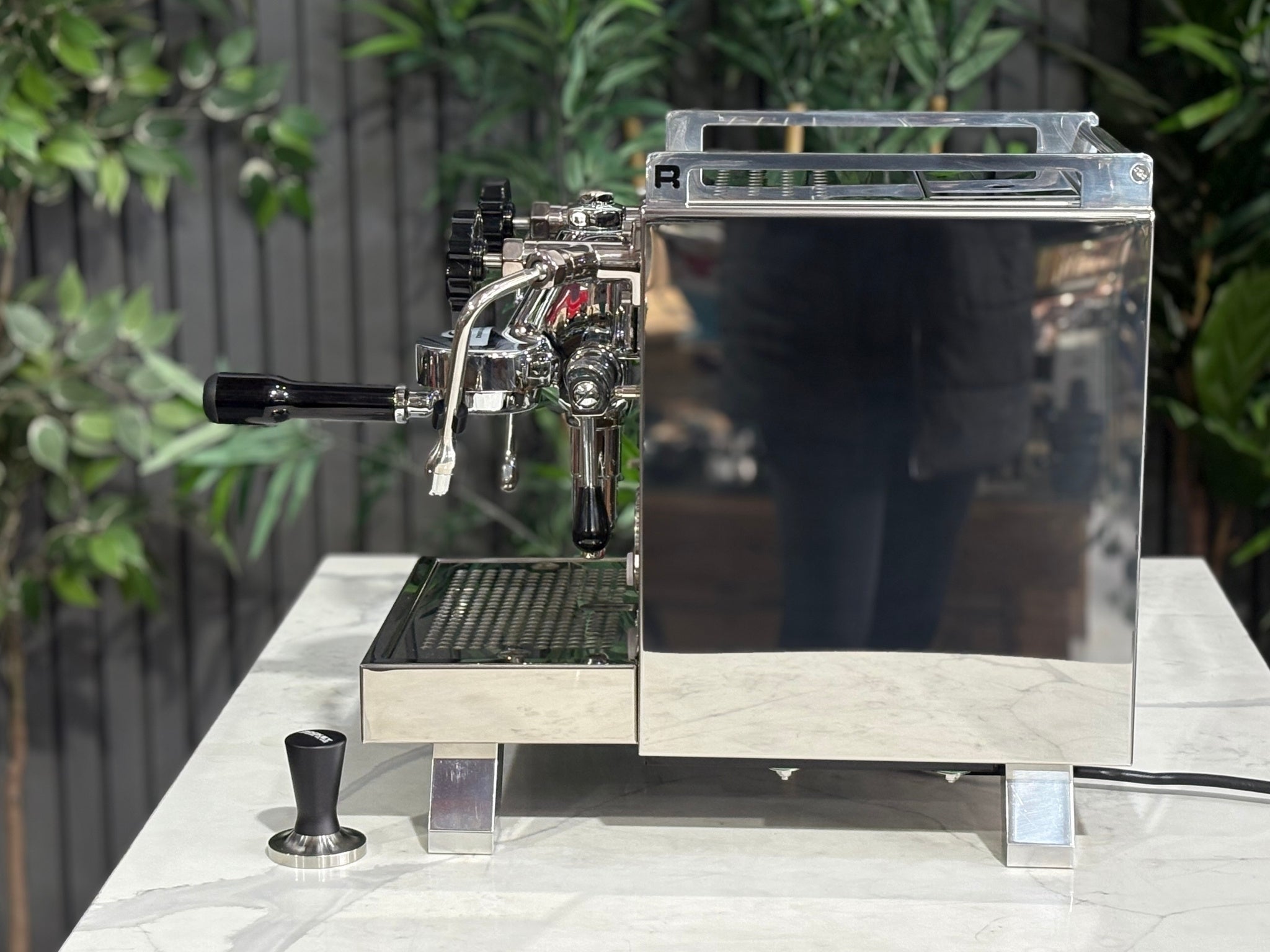 Rocket Cinquantotto R58 1 Group Coffee Machine Stainless - New side profile showing the stainless steel casing, rounded edges, and placement of the steam wand and drip tray on a marble bench with greenery in the background.