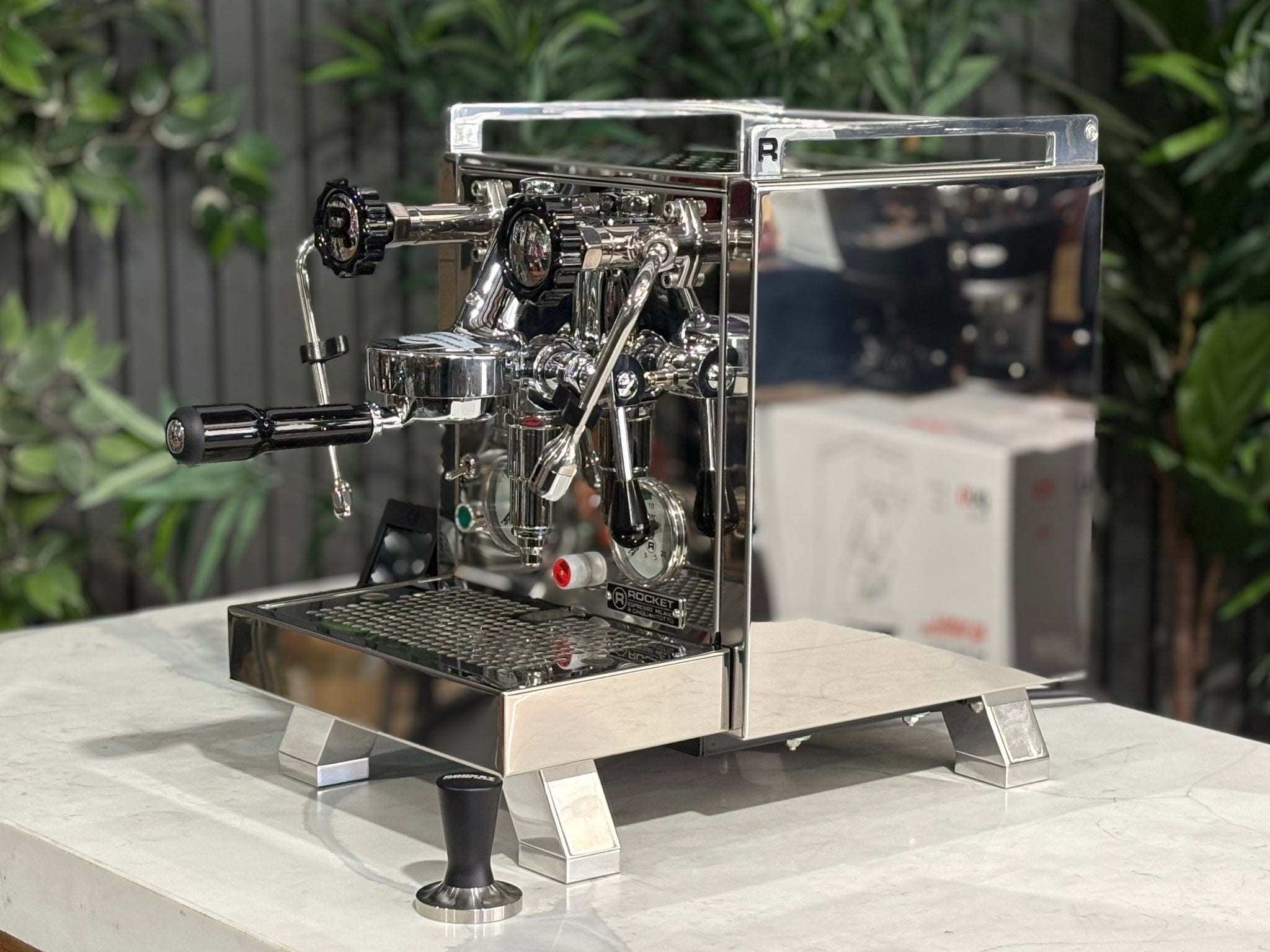 Rocket Cinquantotto R58 1 Group Coffee Machine Stainless - New angled view showing the polished stainless steel body, E61 group head, cool touch steam wand, and wide drip tray on a marble bench with greenery in the background.