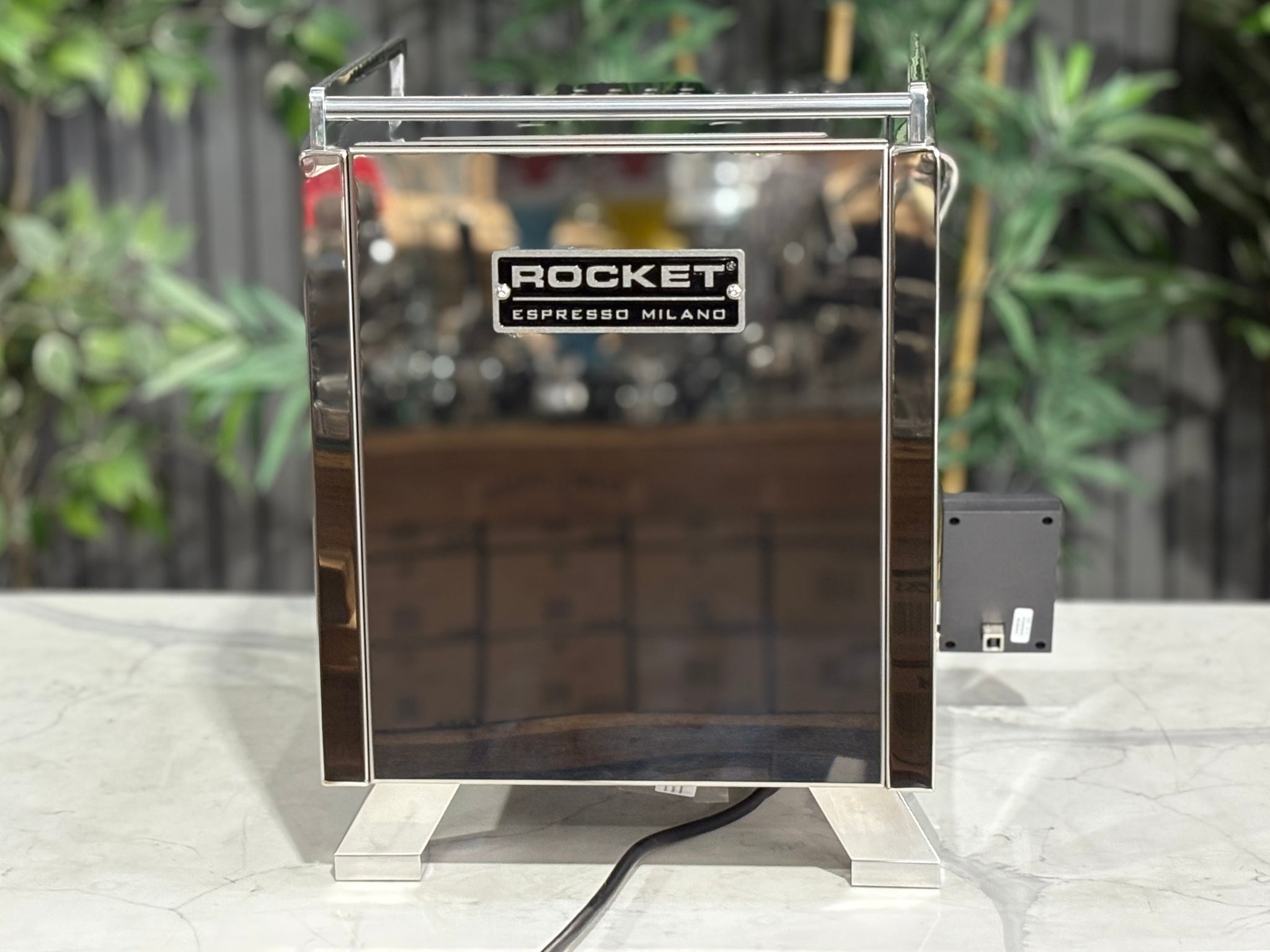 Rocket Cinquantotto R58 1 Group Coffee Machine Stainless - New rear view showing the vertical stainless steel panels, water connection options, and compact footprint on a marble bench with greenery behind.