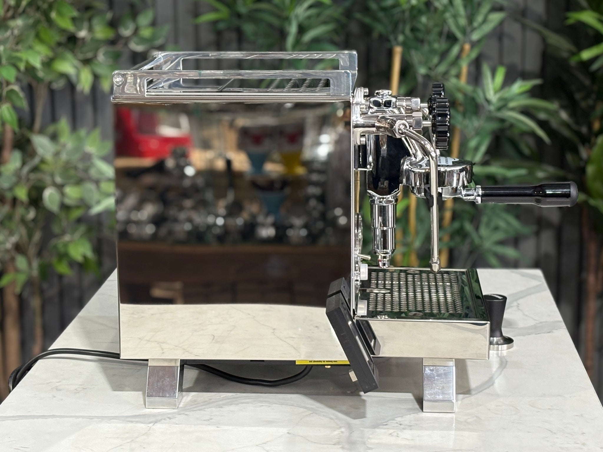 Rocket Cinquantotto R58 1 Group Coffee Machine Stainless - New side profile showing the stainless steel casing, rounded edges, and placement of the steam wand and drip tray on a marble bench with greenery in the background.