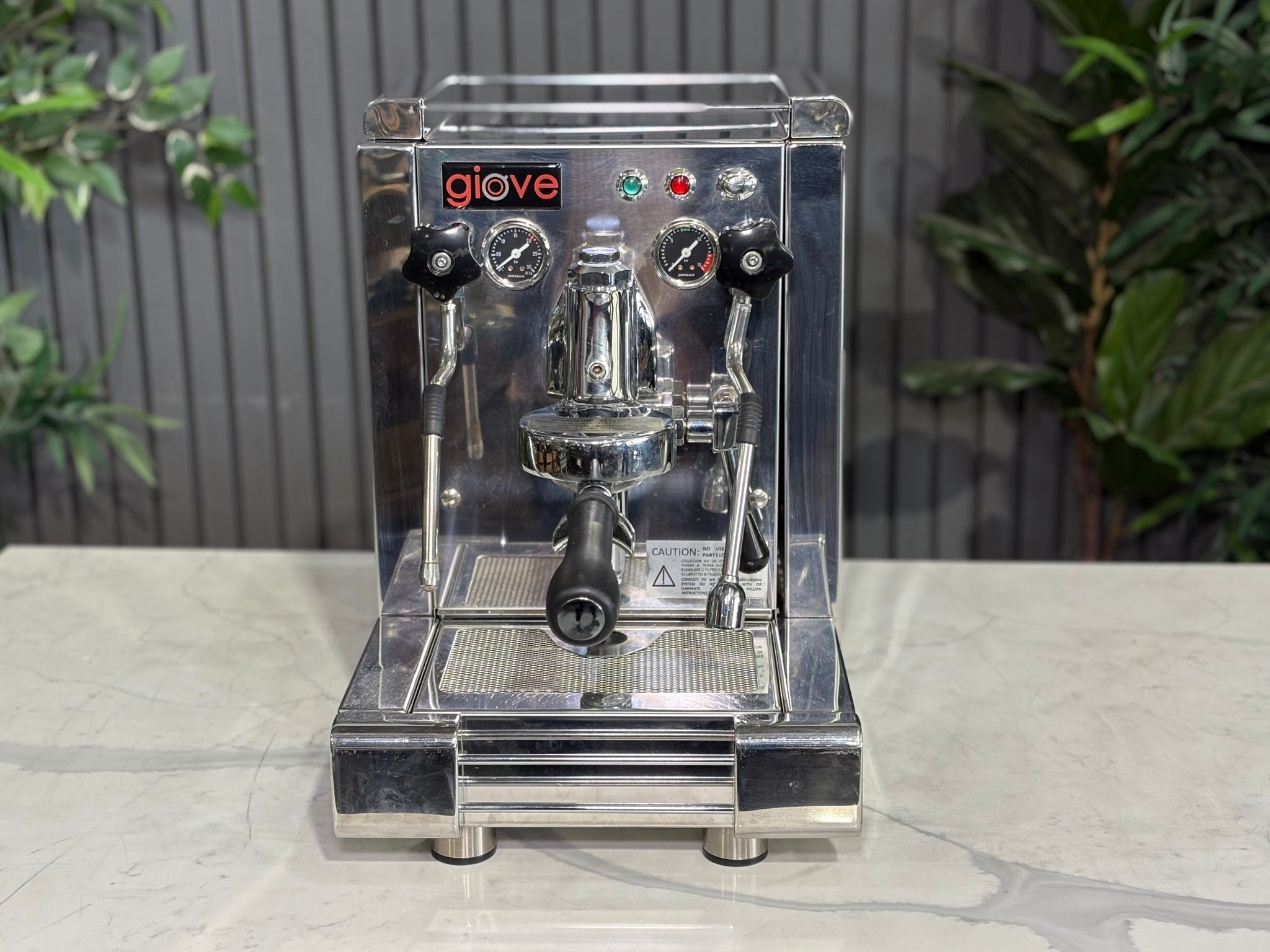 A Royal First Giove 1 Group Espresso Coffee Machine with Stainless finish sitting on a marble bench. There is greenery in the background. The image is taken from the front of the machine so that you can see the front of the machine, control panel, buttons and group head.