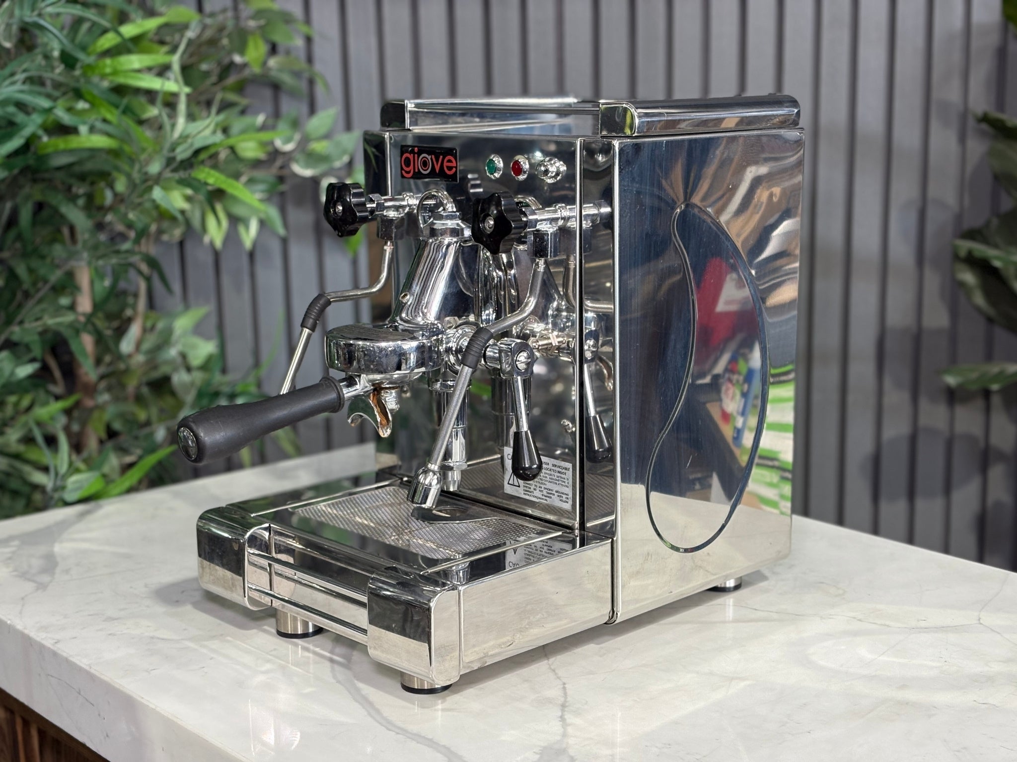 A Royal First Giove 1 Group Espresso Coffee Machine with Stainless finish is sitting on a marble bench. There is greenery in the background. The image is taken from the front of the machine and the machine is sitting on an angle so that you can see the front of the machine and the side.