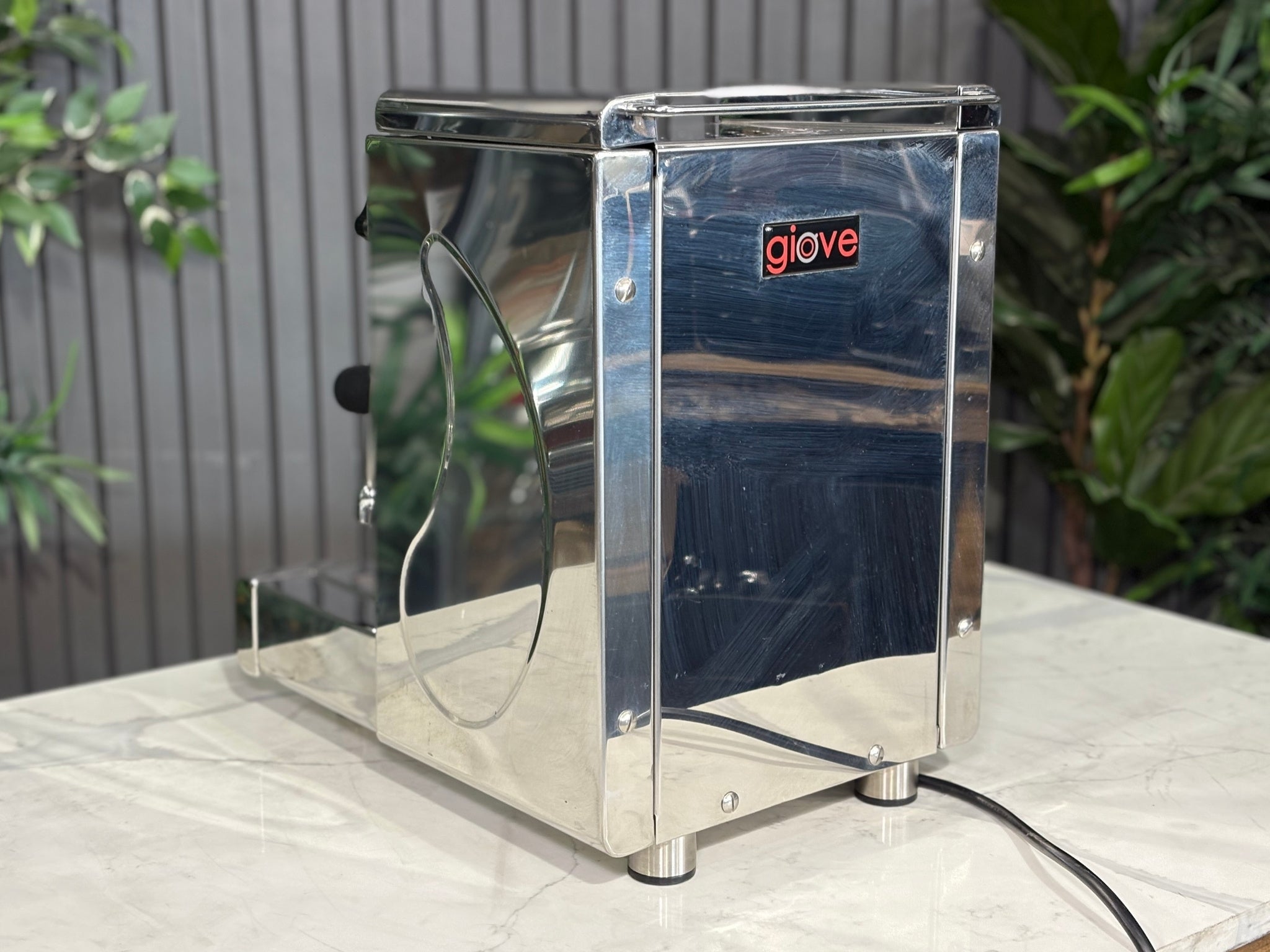 A Royal First Giove 1 Group Espresso Coffee Machine with Stainless finish sitting on a marble bench. There is greenery in the background. The image is taken from behind the machine and the machine is sitting on an angle so that you can see the back of the machine and the side.