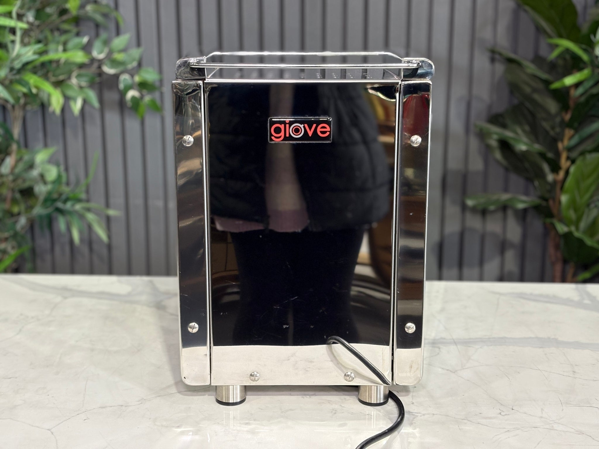 A Royal First Giove 1 Group Espresso Coffee Machine with Stainless finish is sitting on a marble bench. There is greenery in the background. The image is taken from behind the machine so the back is visible. The back of the machine is a stainless steel panel with a red Giove logo in the middle at the top of the machine.