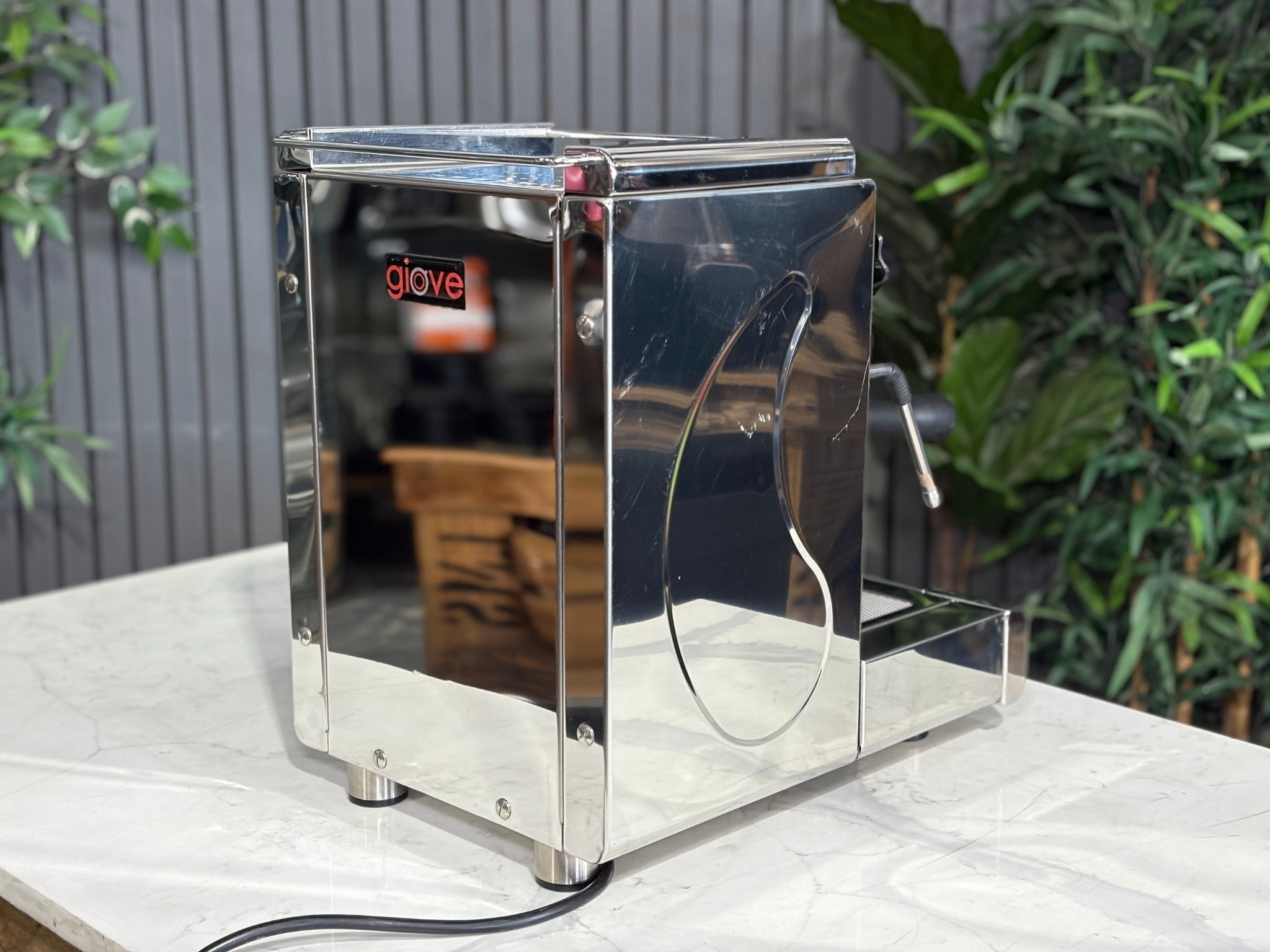 A Royal First Giove 1 Group Espresso Coffee Machine with Stainless finish sitting on a marble bench. There is greenery in the background. The image is taken from behind the machine and the machine is sitting on an angle so that you can see the back of the machine and the side.