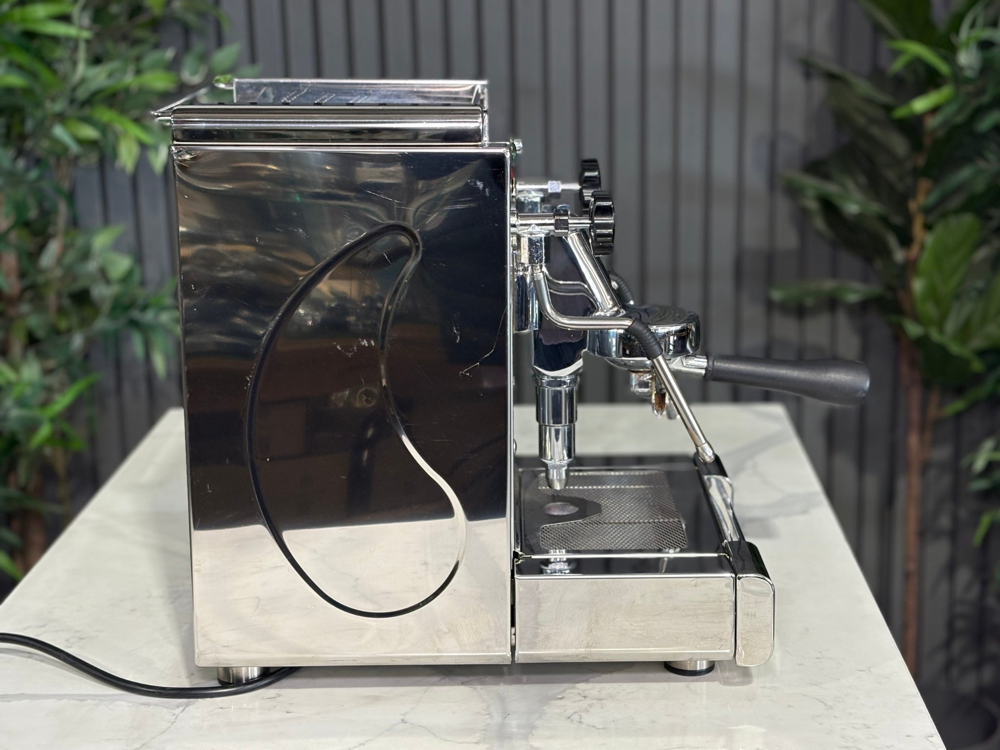 A Royal First Giove 1 Group Espresso Coffee Machine with Stainless finish sitting on a marble bench. There is greenery in the background. The image is taken from the side of the machine.