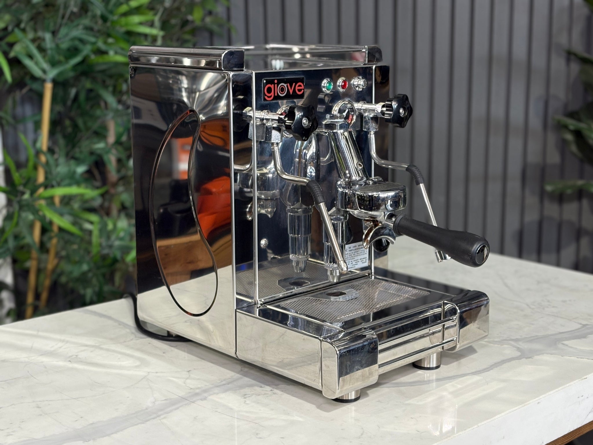 A Royal First Giove 1 Group Espresso Coffee Machine with Stainless finish is sitting on a marble bench. There is greenery in the background. The image is taken from the front of the machine and the machine is sitting on an angle so that you can see the front of the machine and the side.