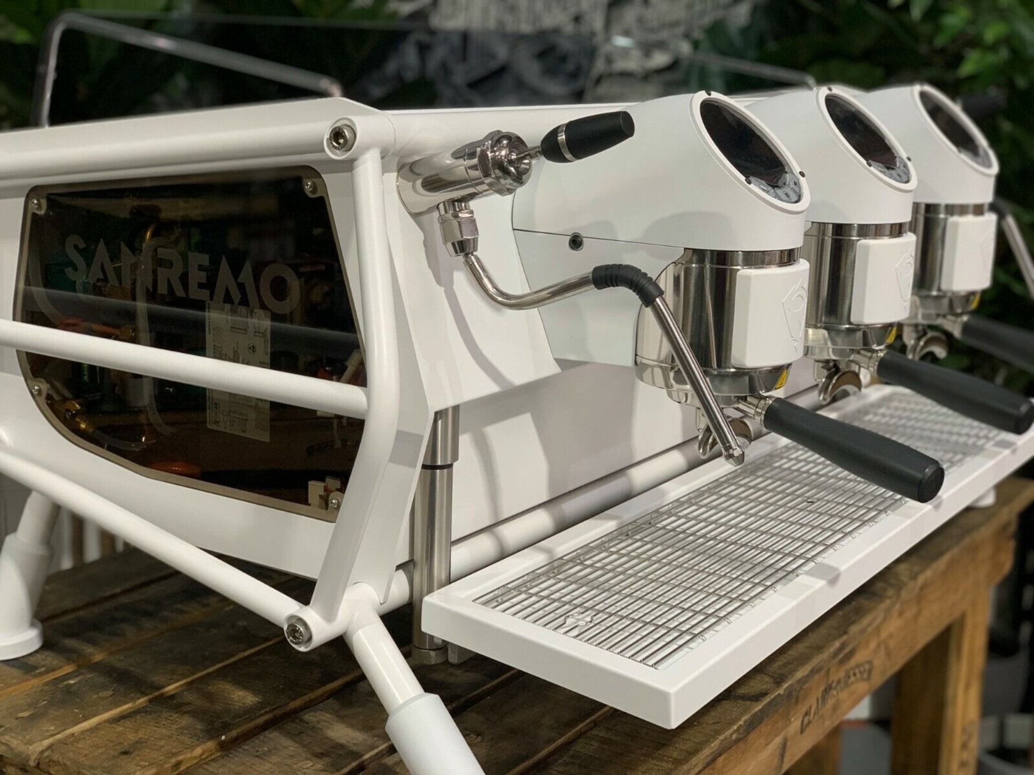 A new San Remo Cafe Racer 3 Group Coffee Machine Naked with white finish is sitting on a wooden bench. There is greenery in the background. The image is a close up image taken from the front of the machine and the machine is sitting on an angle so that you can see the front of the machine, the controls including buttons, group handles and logo.