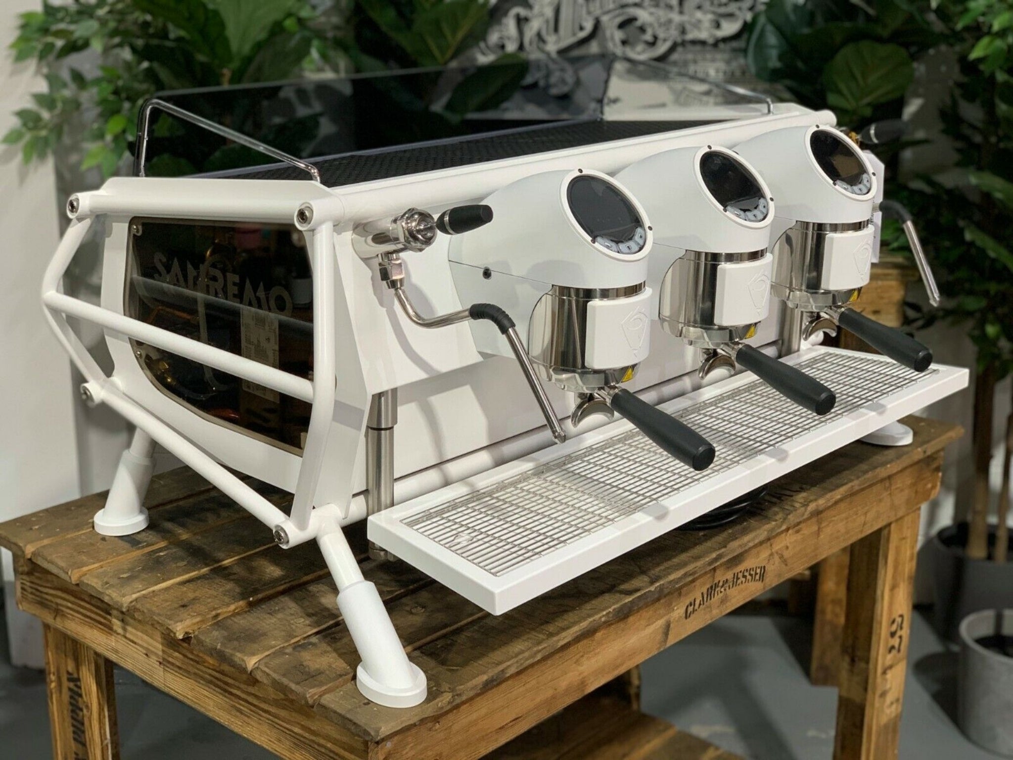 A new San Remo Cafe Racer 3 Group Coffee Machine Naked with white finish
is sitting on a wooden bench. There is greenery in the background. The image is taken from the front of the machine and the machine is sitting on an angle so that you can see the front of the machine and the side.