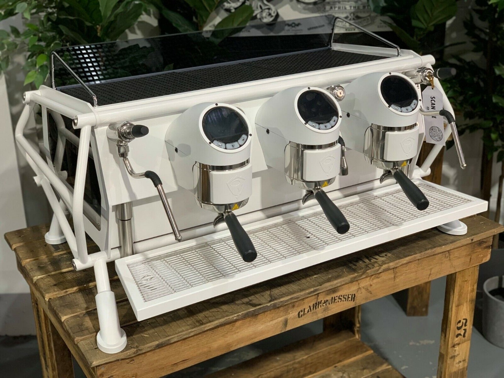 A new San Remo Cafe Racer 3 Group Coffee Machine Naked with white finish
is sitting on a wooden bench. There is greenery in the background. The image is taken from the front of the machine and the machine is sitting on an angle so that you can see the front of the machine and the side.