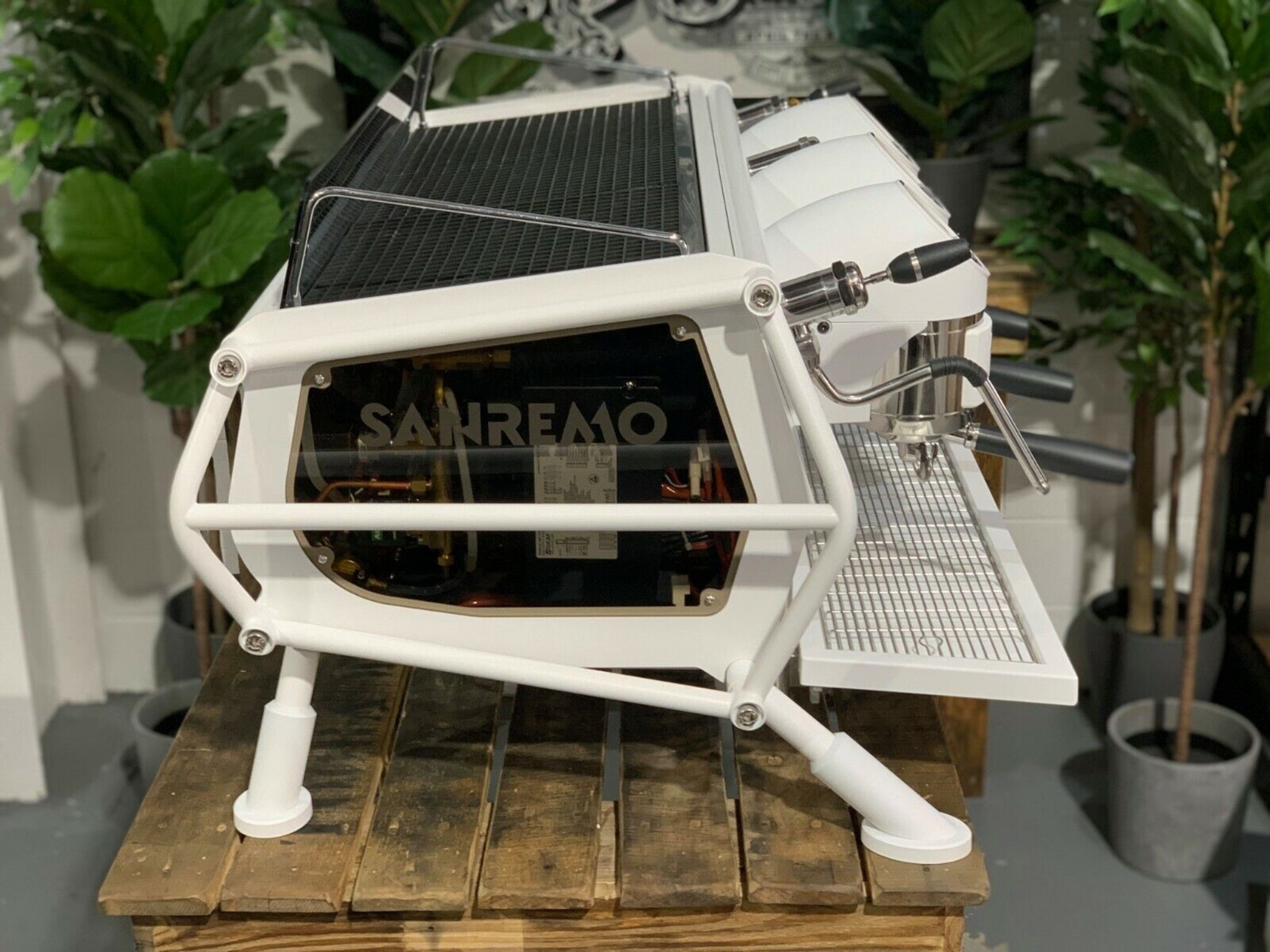 A new San Remo Cafe Racer 3 Group Coffee Machine Naked with white finish sitting on a wooden bench. There is greenery in the background. The image is taken from the side of the machine.