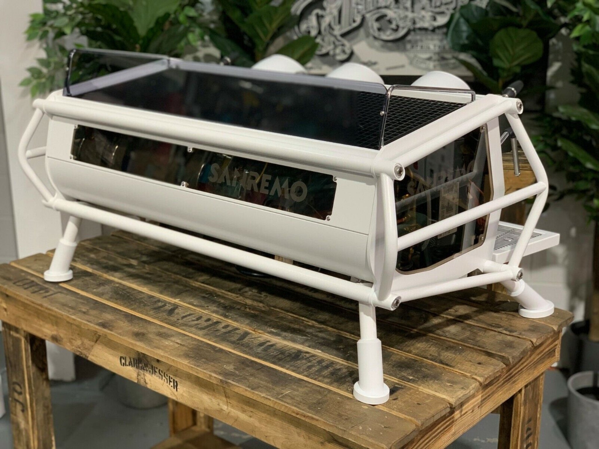 A new San Remo Cafe Racer 3 Group Coffee Machine Naked with white finish  sitting on a wooden bench. There is greenery in the background. The image is taken from behind the machine and the machine is sitting on an angle so that you can see the back of the machine and the side.