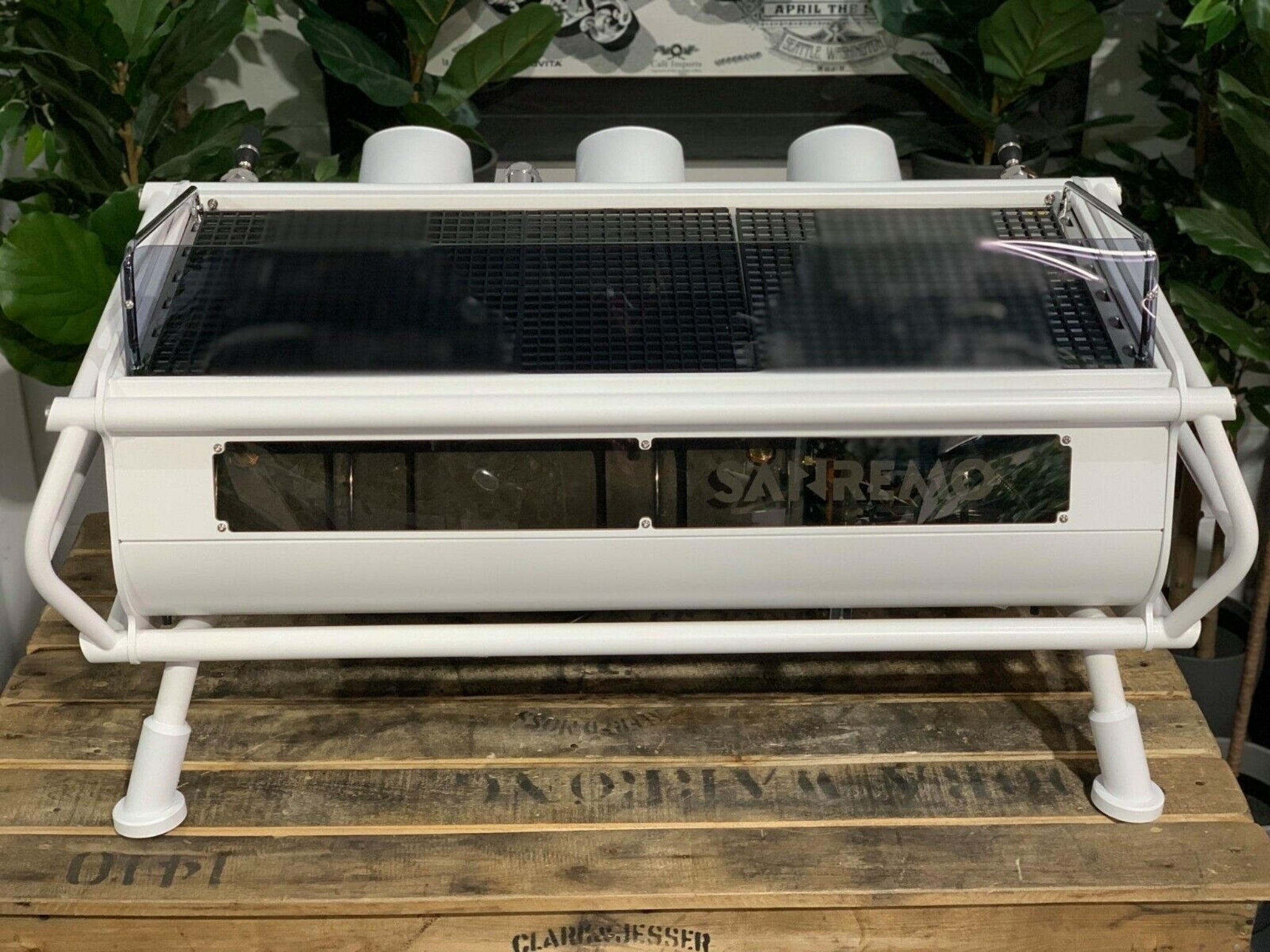 A new San Remo Cafe Racer 3 Group Coffee Machine Naked with white finish
is sitting on a wooden bench. There is greenery in the background. The image is taken from behind the machine so the back is visible.