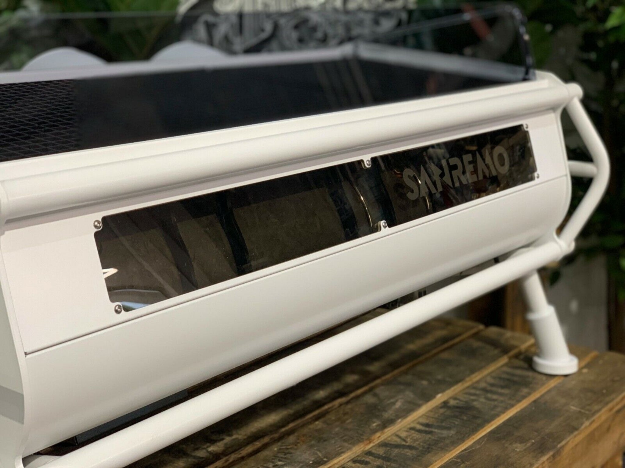 A new San Remo Cafe Racer 3 Group Coffee Machine Naked with white finish
is sitting on a wooden bench. There is greenery in the background. The image is a close up image taken from the back of the machine and the machine is sitting on an angle so that you can see the back of the machine, with its clear panel and Sanremo logo.