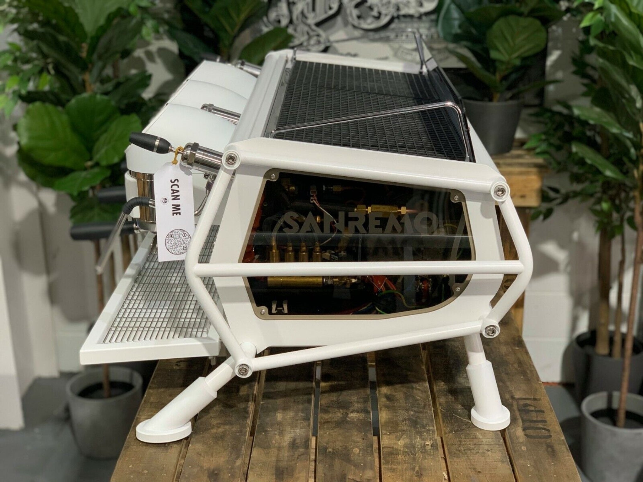 A new San Remo Cafe Racer 3 Group Coffee Machine Naked with white finish sitting on a wooden bench. There is greenery in the background. The image is taken from the side of the machine.