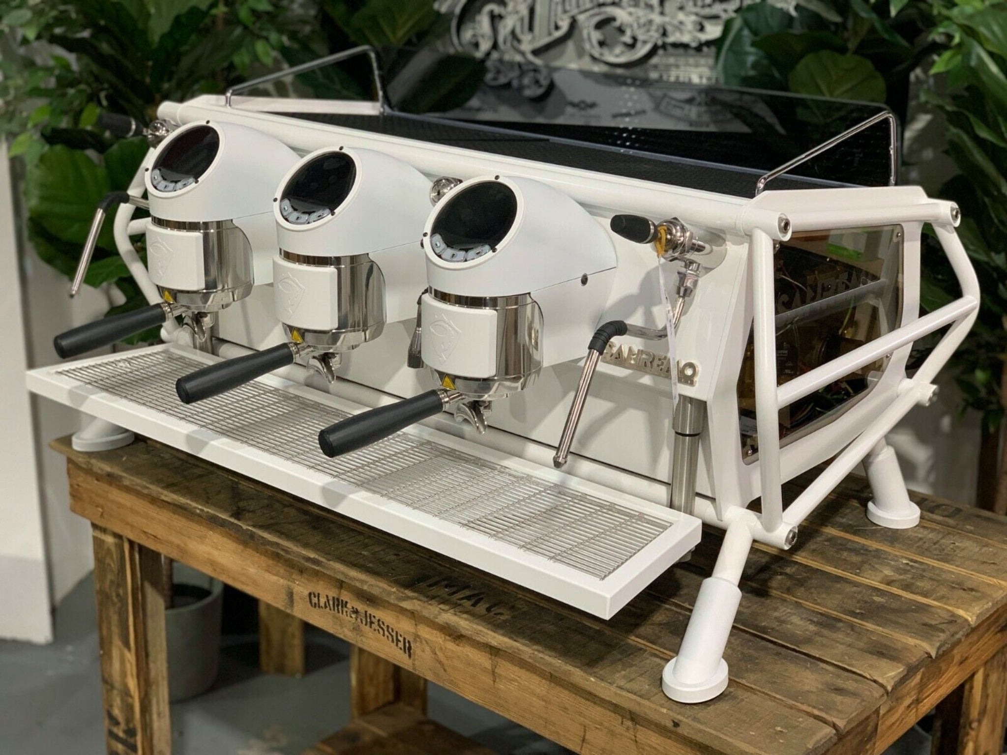 A new San Remo Cafe Racer 3 Group Coffee Machine Naked with white finish
is sitting on a wooden bench. There is greenery in the background. The image is taken from the front of the machine and the machine is sitting on an angle so that you can see the front of the machine and the side.