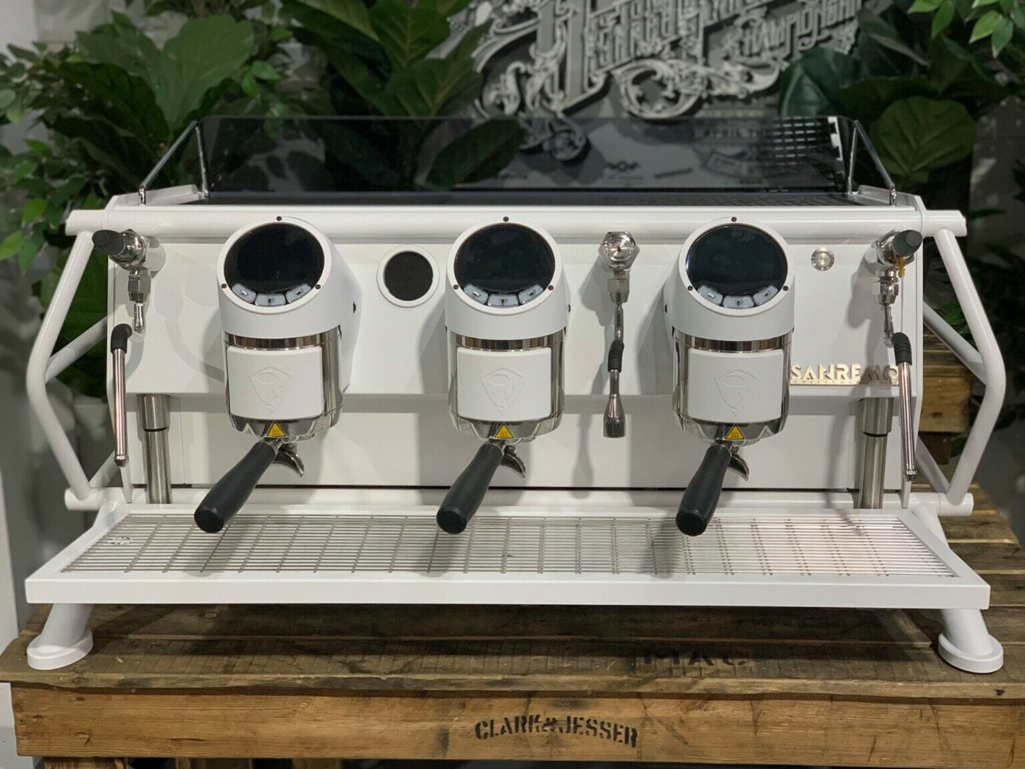 A new San Remo Cafe Racer 3 Group Coffee Machine Naked with white finish sitting on a wooden bench. There is greenery in the background. The image is taken from the front of the machine so that you can see the front of the machine, control panel, buttons and group heads.