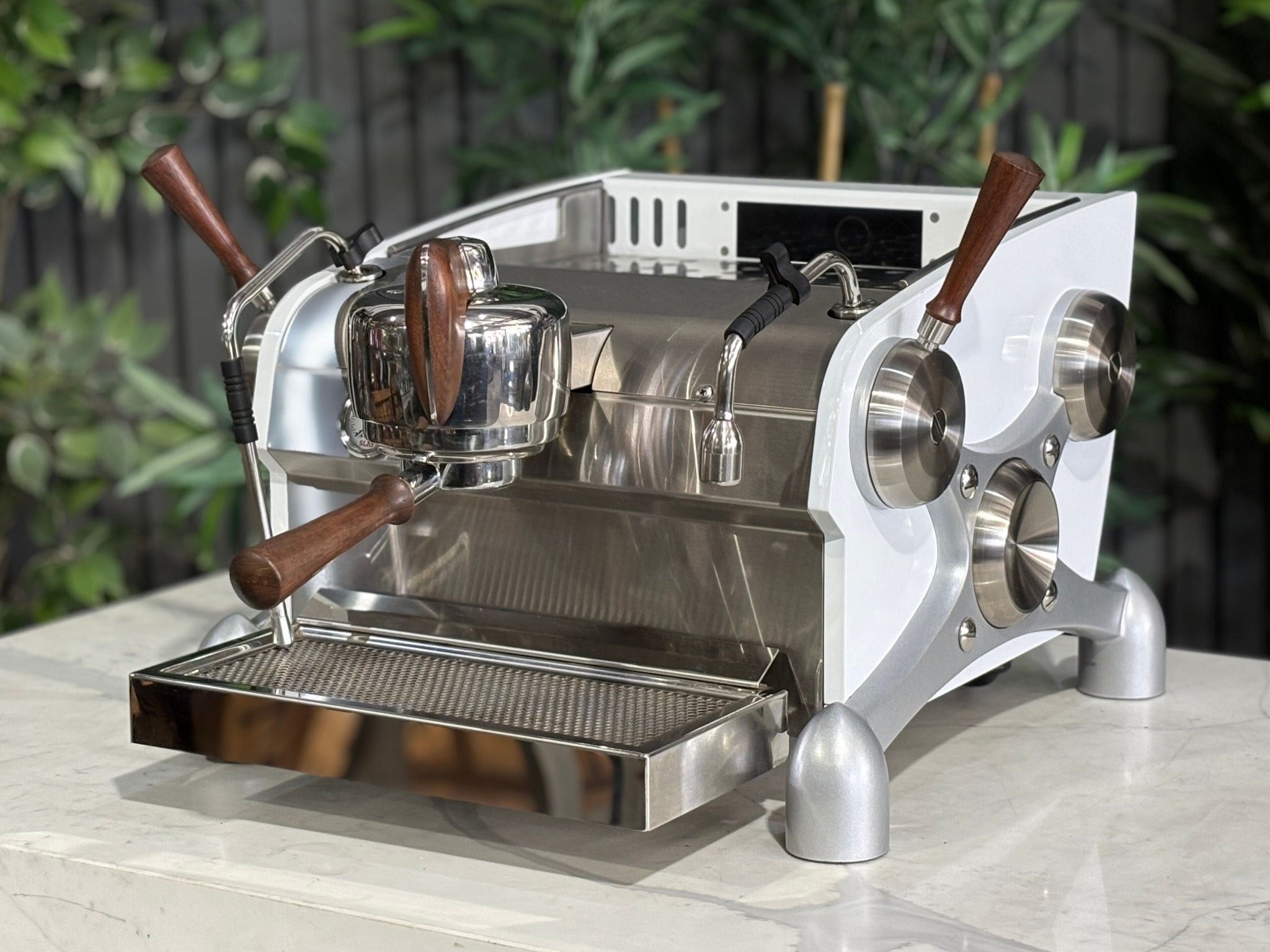 Slayer Espresso 1 Group Coffee Machine White – Used, angled view showing the front and side of the espresso coffee machine with white panels, timber accents, manual paddle, and dual steam wands on a marble bench with greenery in the background.