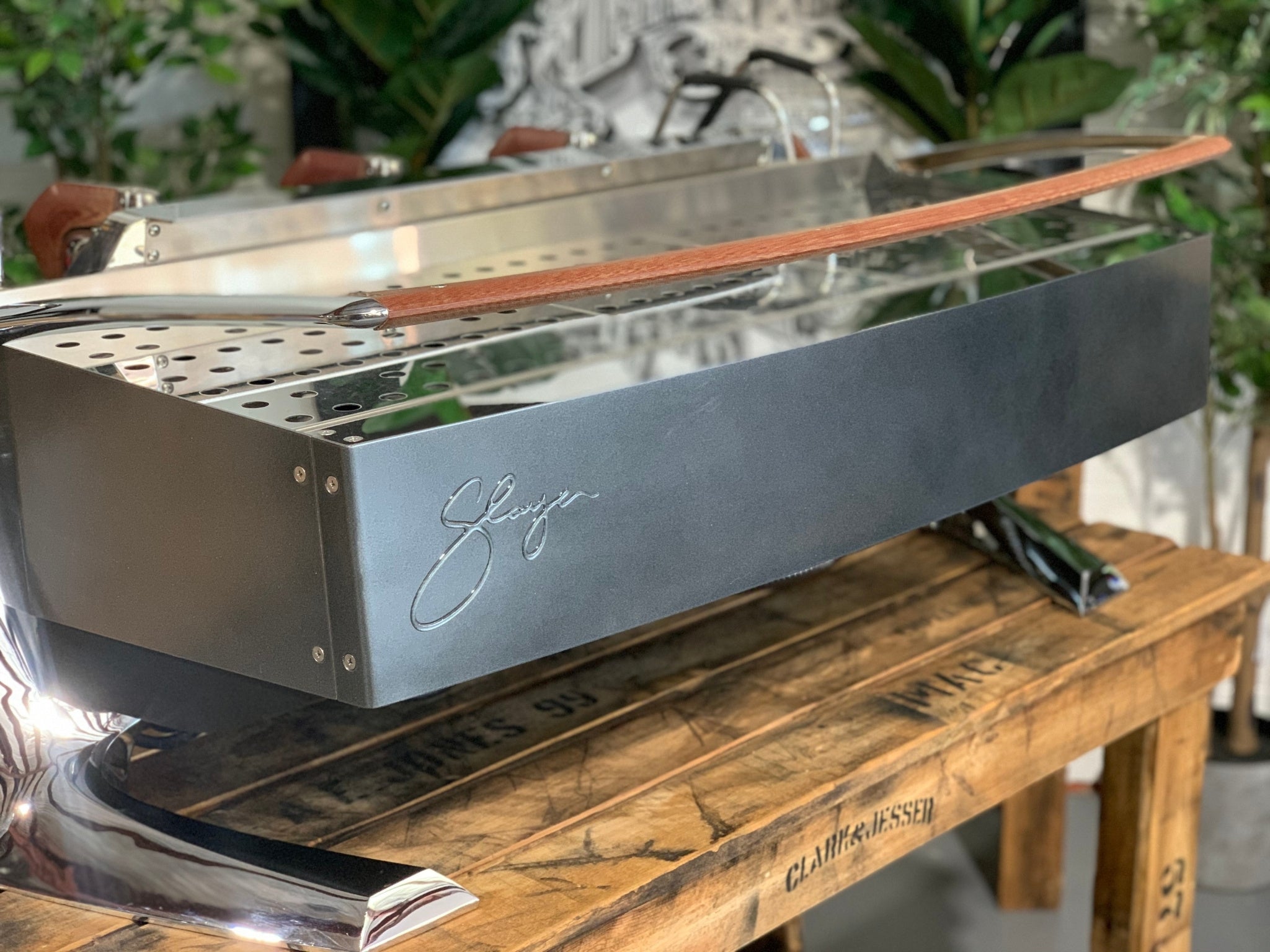 Slayer Steam X 3 Group Espresso Coffee Machine in black sitting on a wooden bench. There is greenery in the background. The image is taken from behind the machine and the machine is sitting on an angle so that you can see the back of the machine and the side.