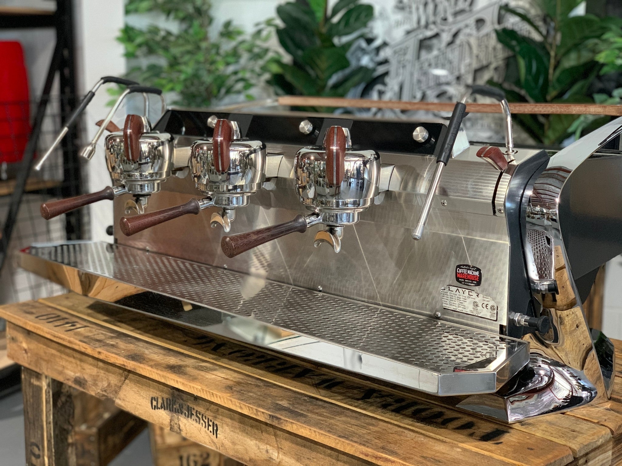 Slayer Steam X 3 Group Espresso Coffee Machine in black is sitting on a wooden bench. There is greenery in the background. The image is taken from the front of the machine and the machine is sitting on an angle so that you can see the front of the machine and the side