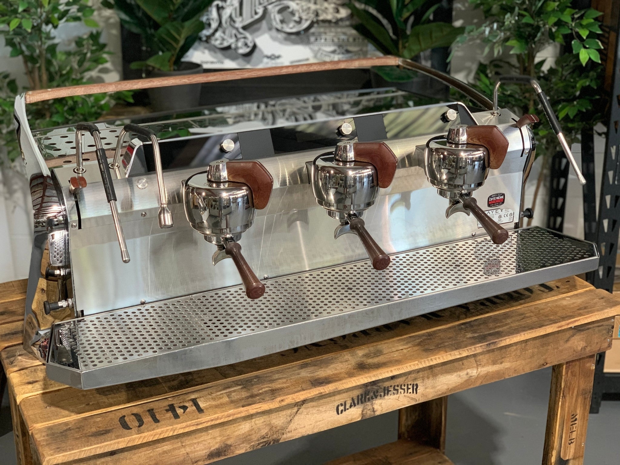 Slayer Steam X 3 Group Espresso Coffee Machine in black is sitting on a wooden bench. There is greenery in the background. The image is taken from the front of the machine and the machine is sitting on an angle so that you can see the front of the machine and the side