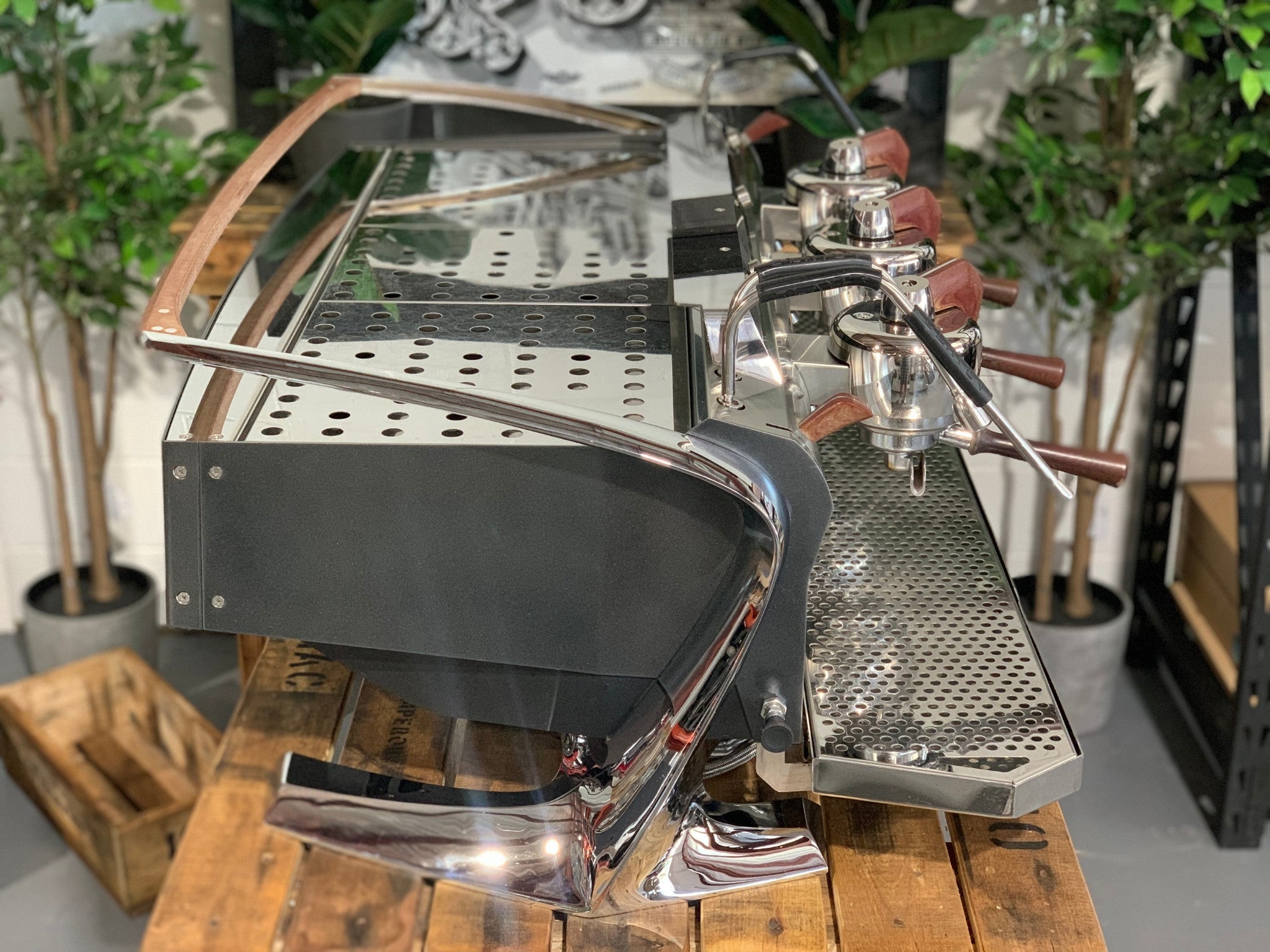 Slayer Steam X 3 Group Espresso Coffee Machine in black sitting on a wooden bench. There is greenery in the background. The image is taken from the side of the machine.