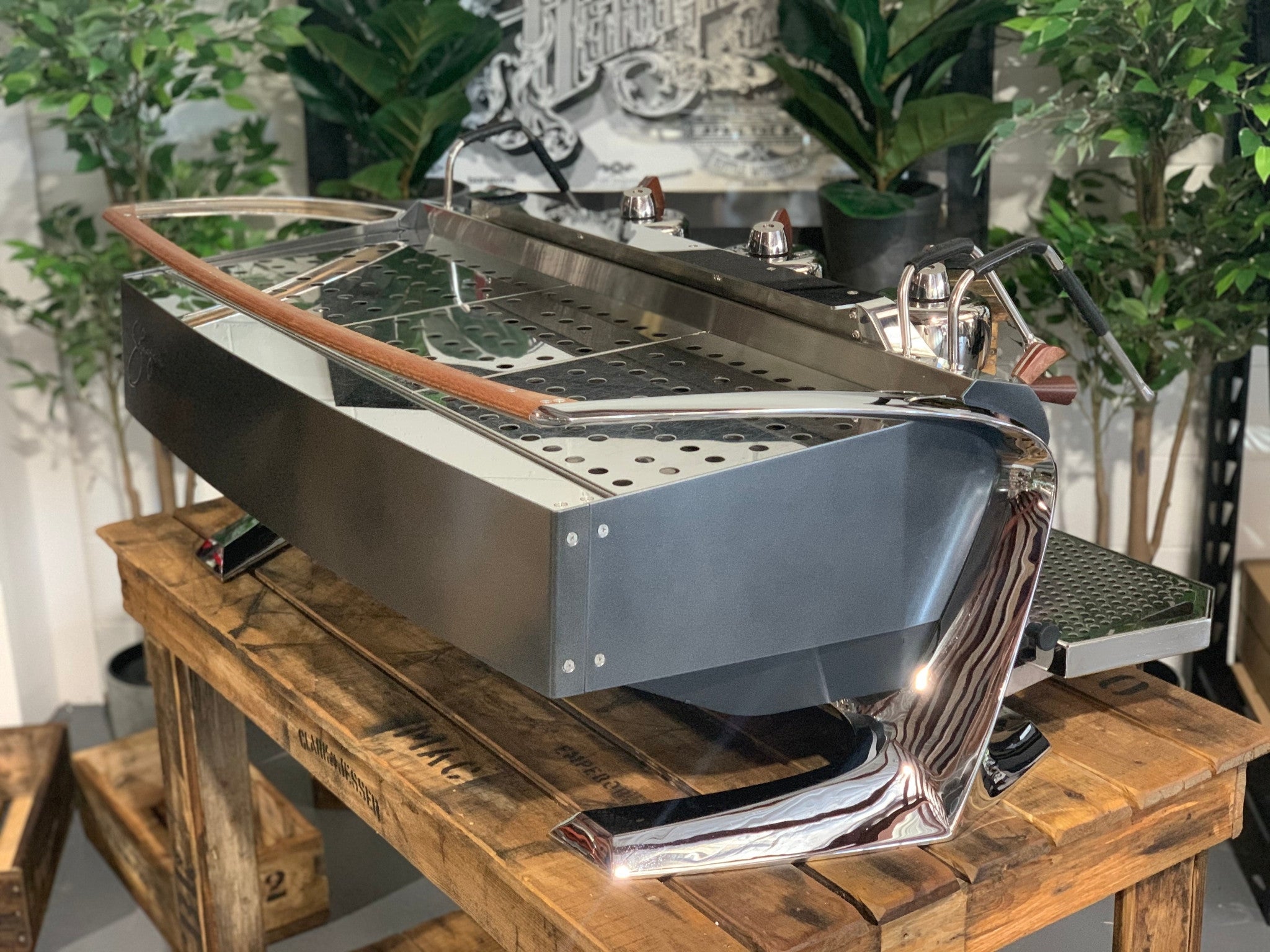 Slayer Steam X 3 Group Espresso Coffee Machine in black sitting on a wooden bench. There is greenery in the background. The image is taken from behind the machine and the machine is sitting on an angle so that you can see the back of the machine and the side.