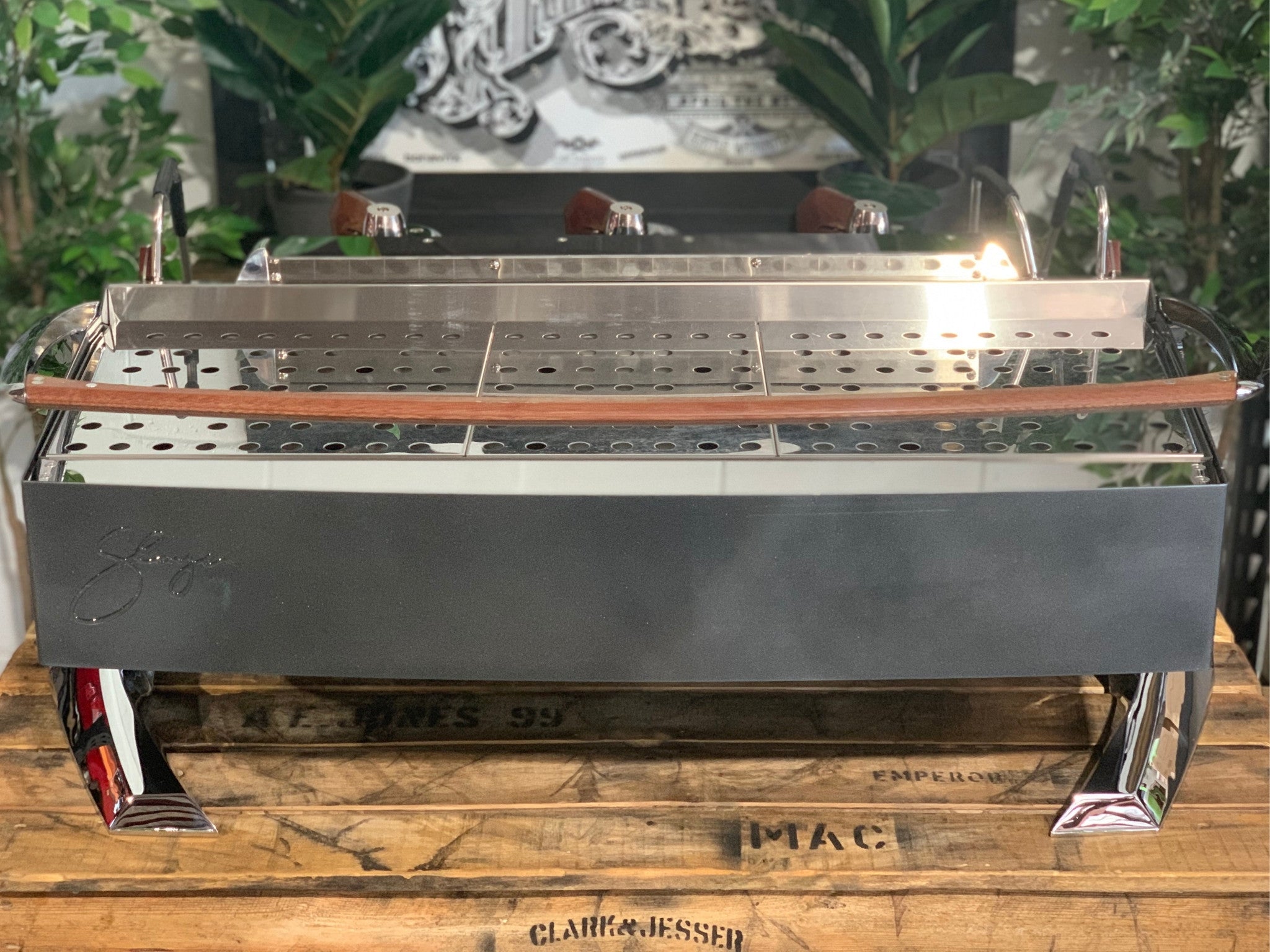 Slayer Steam X 3 Group Espresso Coffee Machine in black is sitting on a wooden bench. There is greenery in the background. The image is taken from behind the machine so the back is visible.