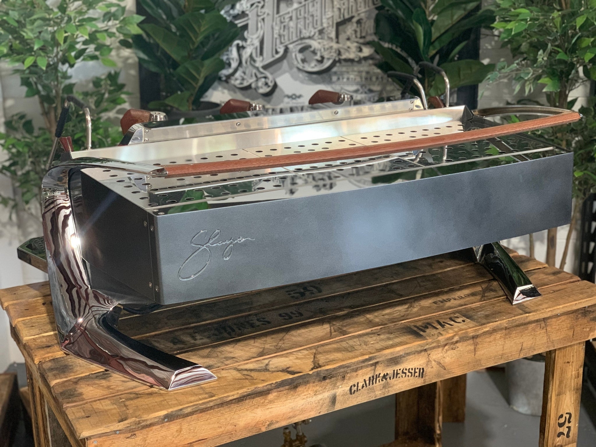 Slayer Steam X 3 Group Espresso Coffee Machine in black sitting on a wooden bench. There is greenery in the background. The image is taken from behind the machine and the machine is sitting on an angle so that you can see the back of the machine and the side.