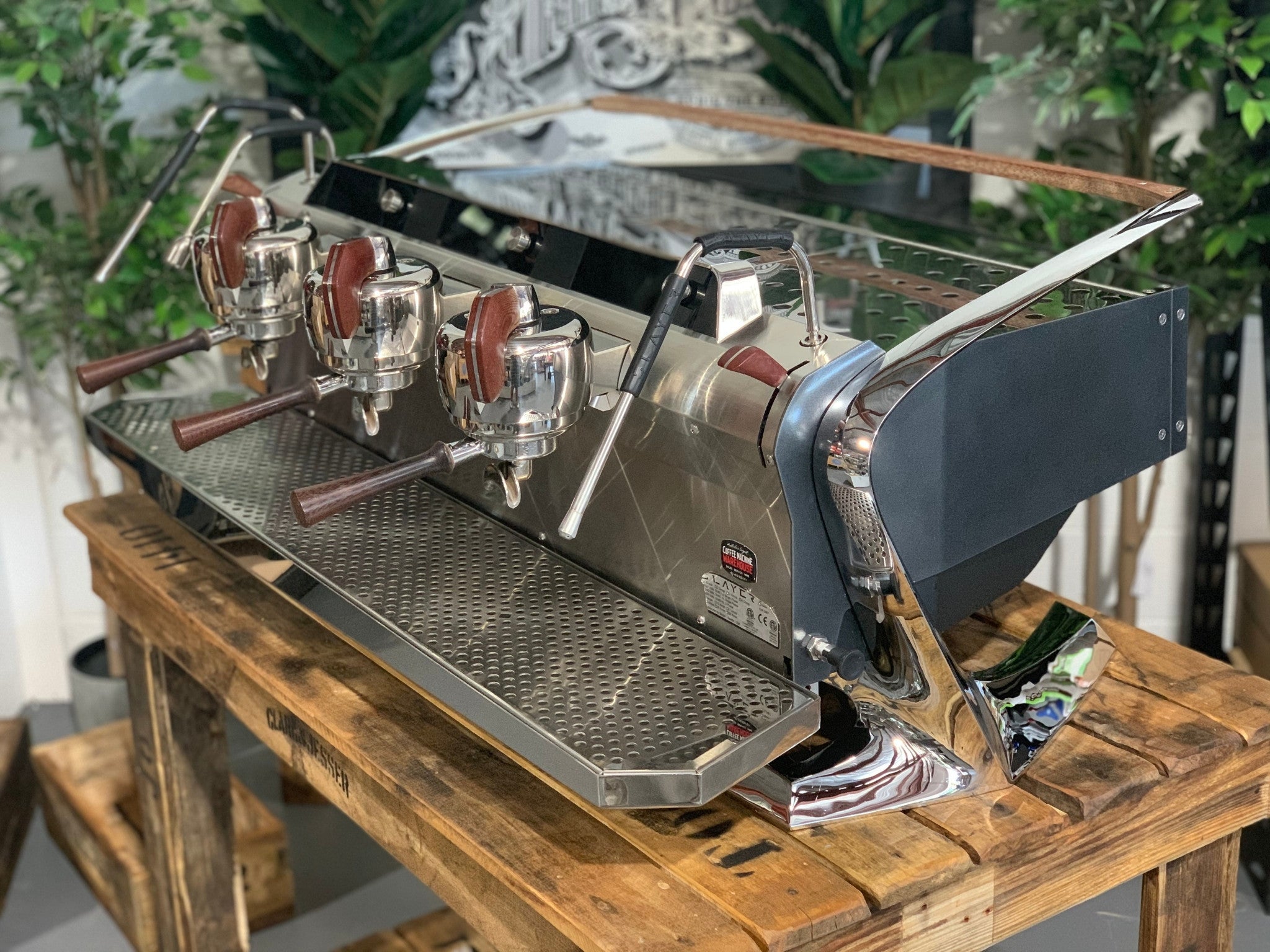 Slayer Steam X 3 Group Espresso Coffee Machine in black  is sitting on a wooden bench. There is greenery in the background. The image is taken from the front of the machine and the machine is sitting on an angle so that you can see the front of the machine and the side.