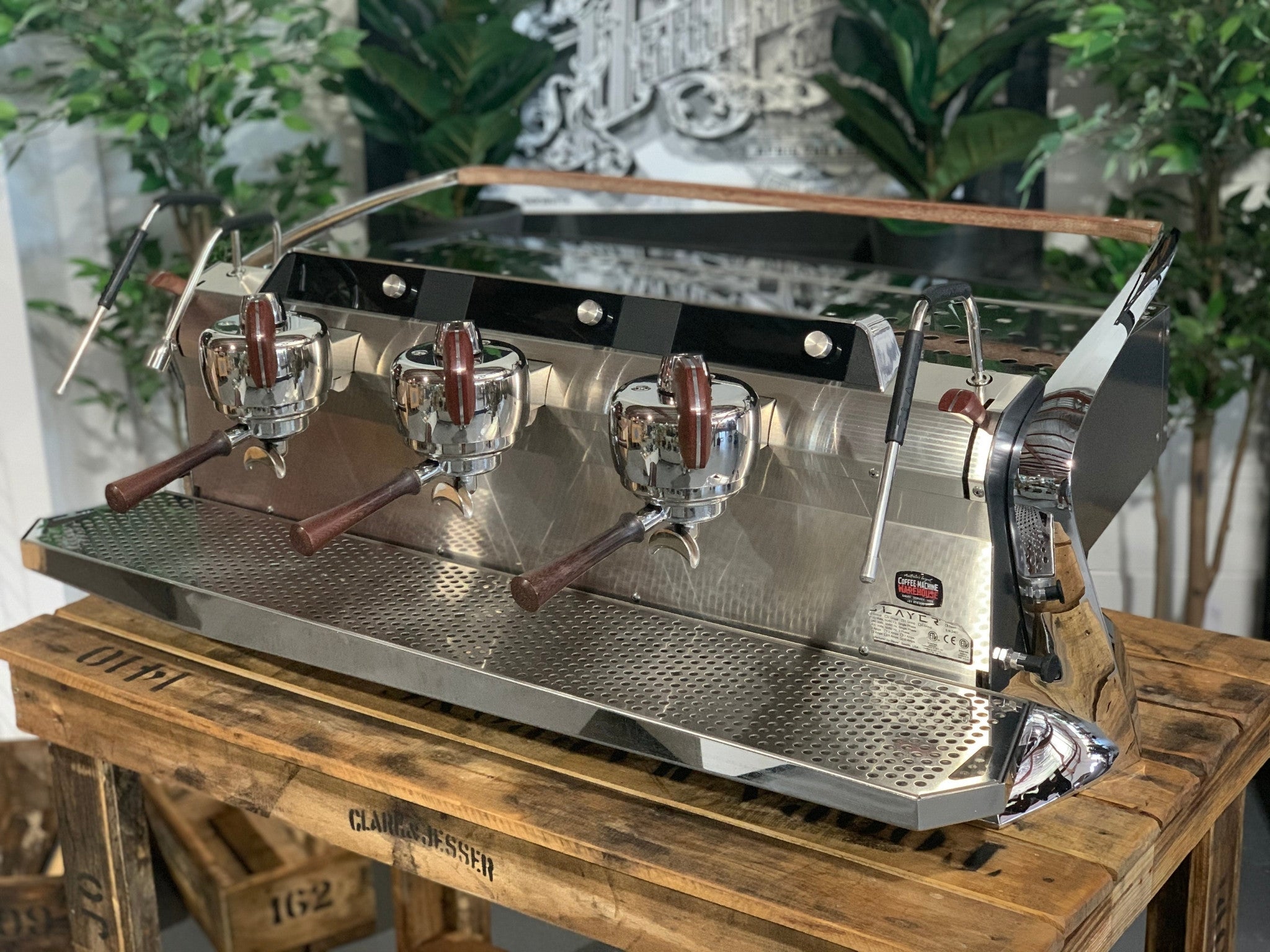 Slayer Steam X 3 Group Espresso Coffee Machine in black
 is sitting on a wooden bench. There is greenery in the background. The image is taken from the front of the machine and the machine is sitting on an angle so that you can see the front of the machine and the side