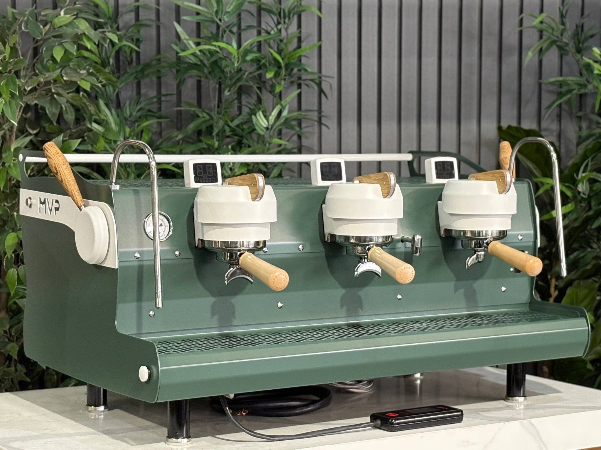 Synesso MVP 3 Group Coffee Machine Green, White & Timber - Used, angled view showing the green and white painted body with timber accents, dual group heads, cool touch steam wands, cup tray, and digital shot timers on a marble bench with greenery in the background.