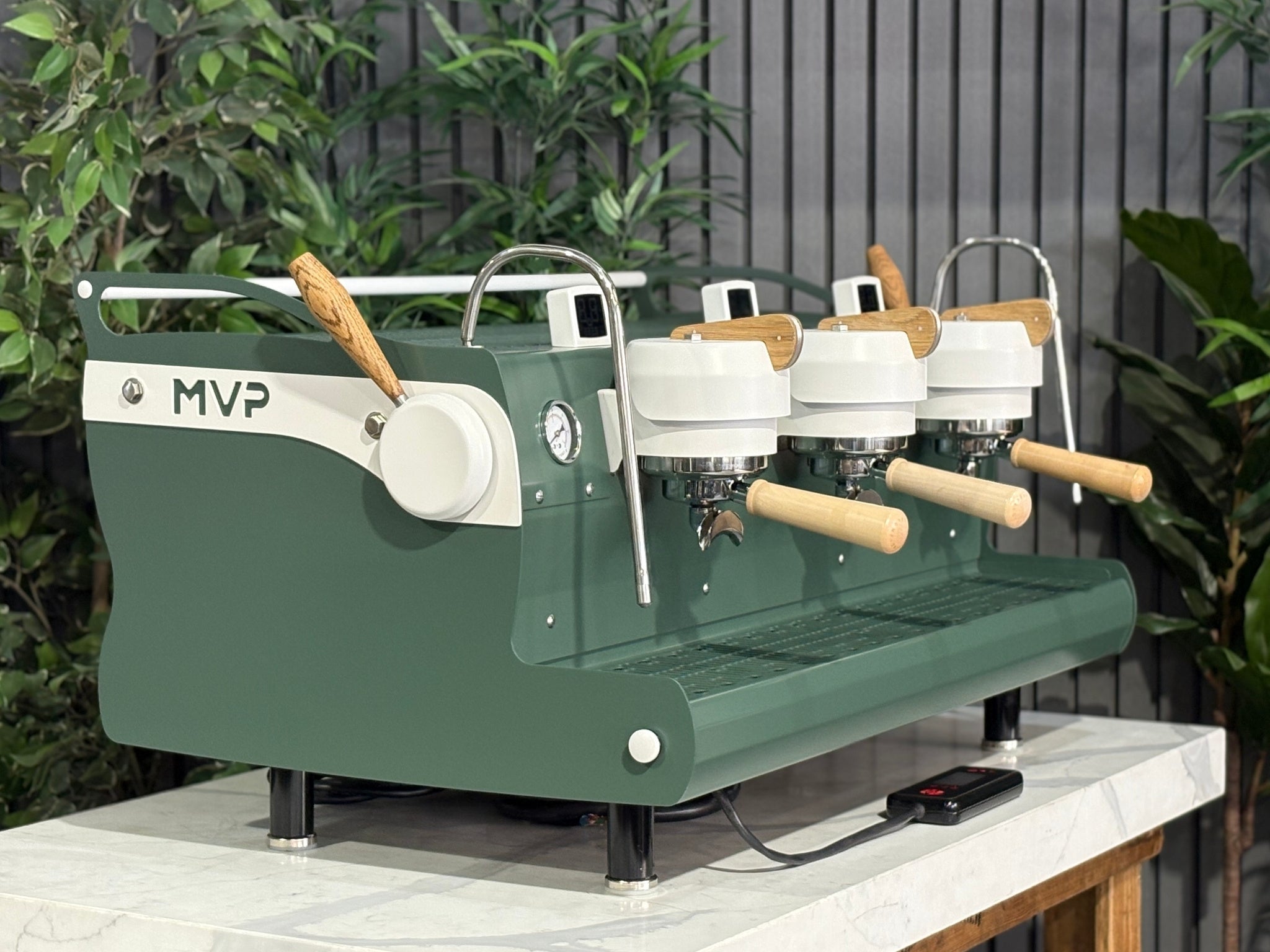 Synesso MVP 3 Group Coffee Machine Green, White & Timber - Used, angled view showing the green and white painted body with timber accents, dual group heads, cool touch steam wands, cup tray, and digital shot timers on a marble bench with greenery in the background.