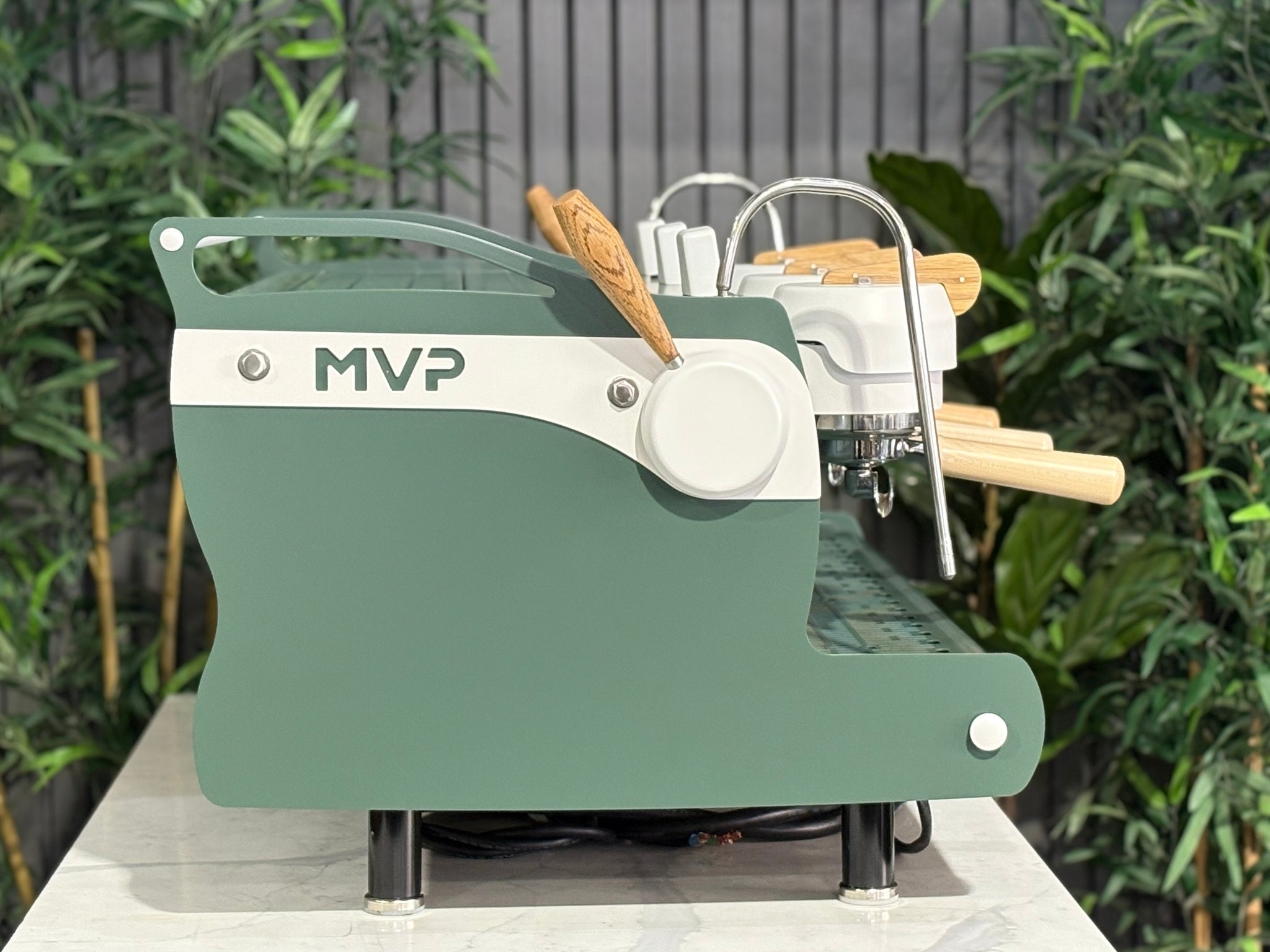 Synesso MVP 3 Group Coffee Machine Green, White & Timber - Used, side view showing the green and white painted body, timber detailing, dual steam wands, and ergonomic lever handles, on a marble bench with greenery behind.