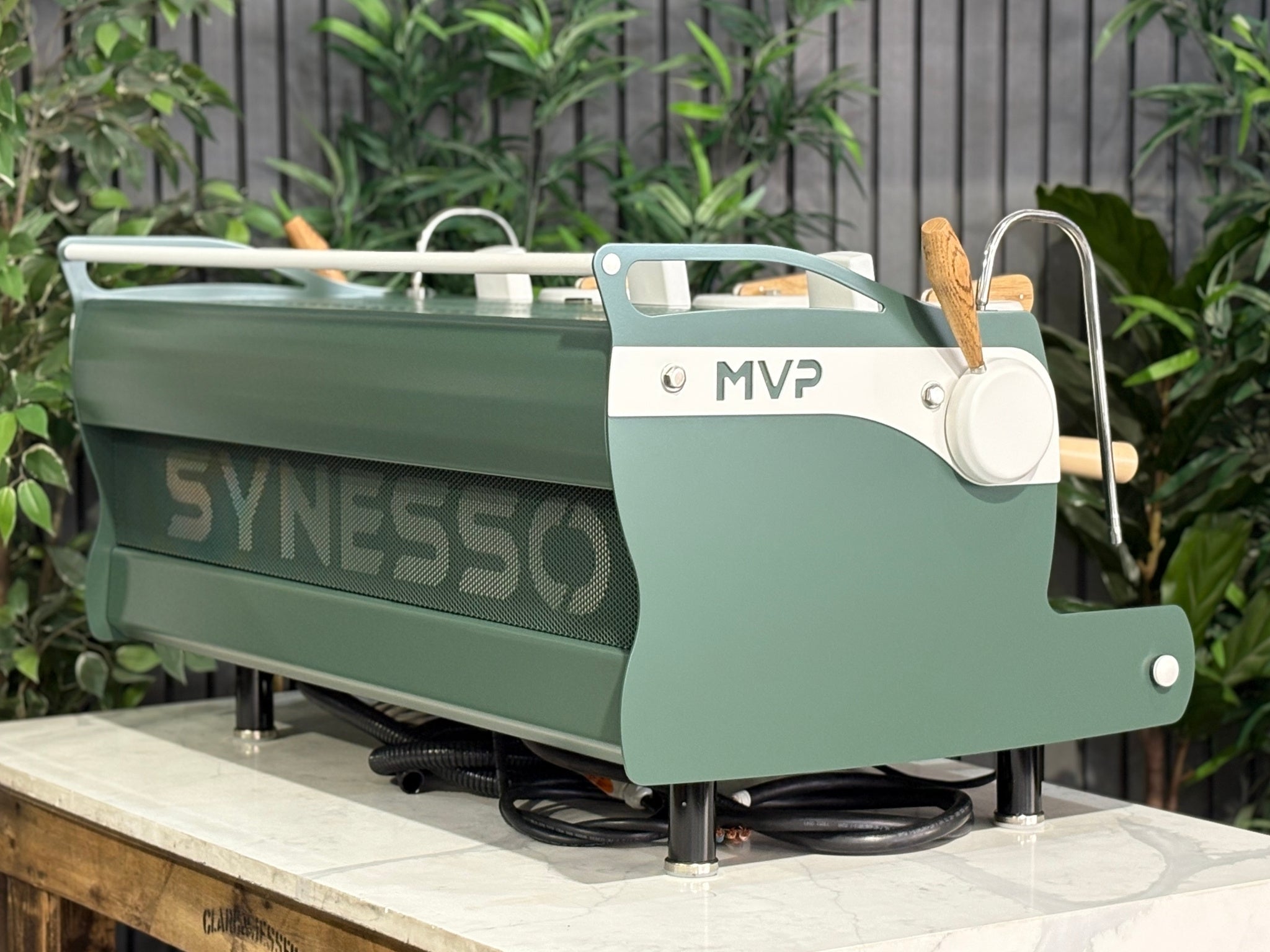 Synesso MVP 3 Group Coffee Machine Green, White & Timber - Used, angled rear view showing stainless steel back panel, green and white painted body, timber accents, and part of the side, set on a marble bench with greenery in the background.