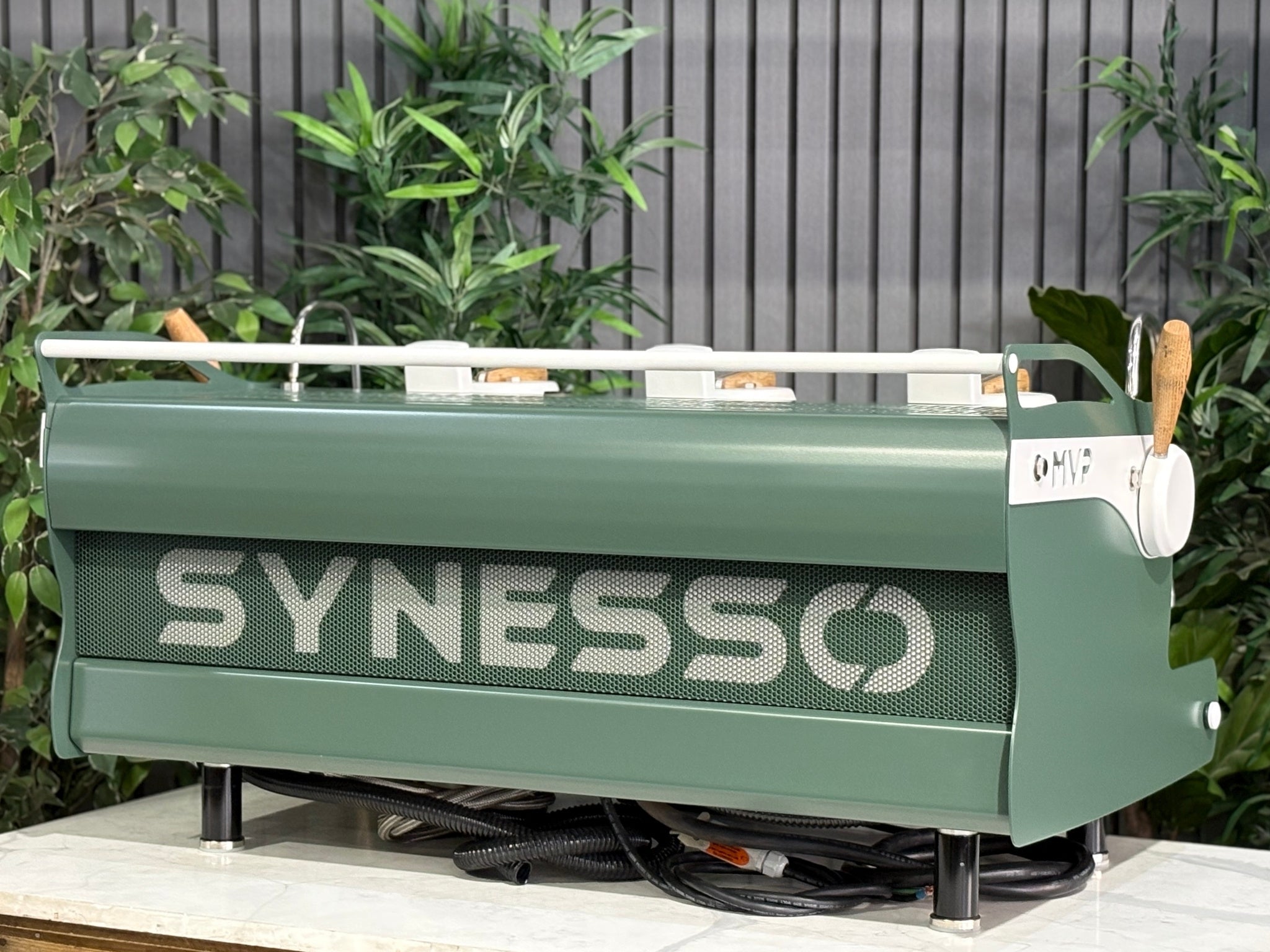 Synesso MVP 3 Group Coffee Machine Green, White & Timber - Used, angled rear view showing stainless steel back panel, green and white painted body, timber accents, and part of the side, set on a marble bench with greenery in the background.