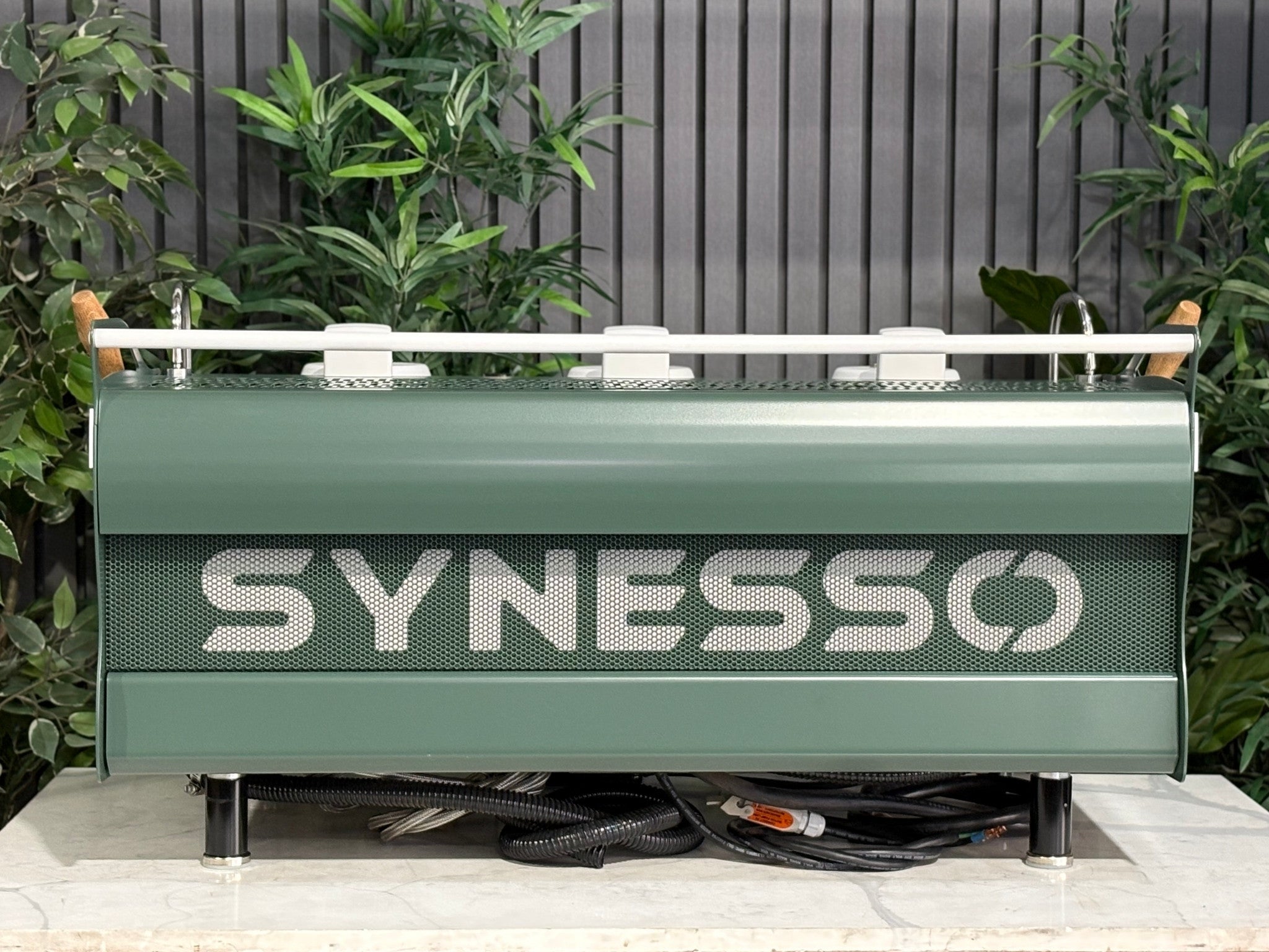 Synesso MVP 3 Group Coffee Machine Green, White & Timber - Used, rear view displaying stainless steel back panel, green and white painted sides, and timber accents, positioned on a marble bench with greenery behind.