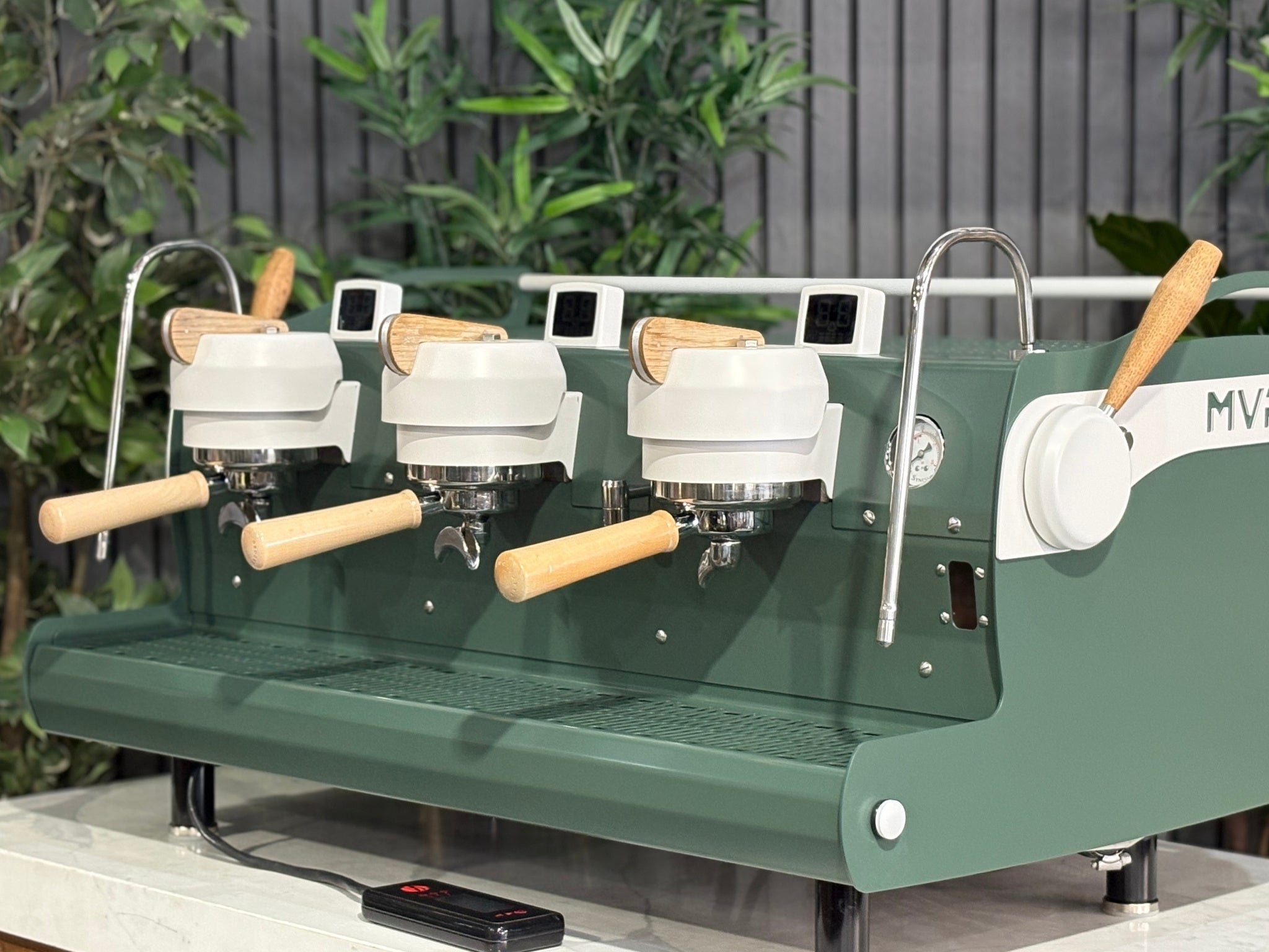 Synesso MVP 3 Group Coffee Machine Green, White & Timber - Used, close-up angled view showing dual group heads, digital shot timers, dual steam wands with ergonomic lever handles, and cup tray, featuring green and white painted body with timber accents on a marble bench with greenery behind.