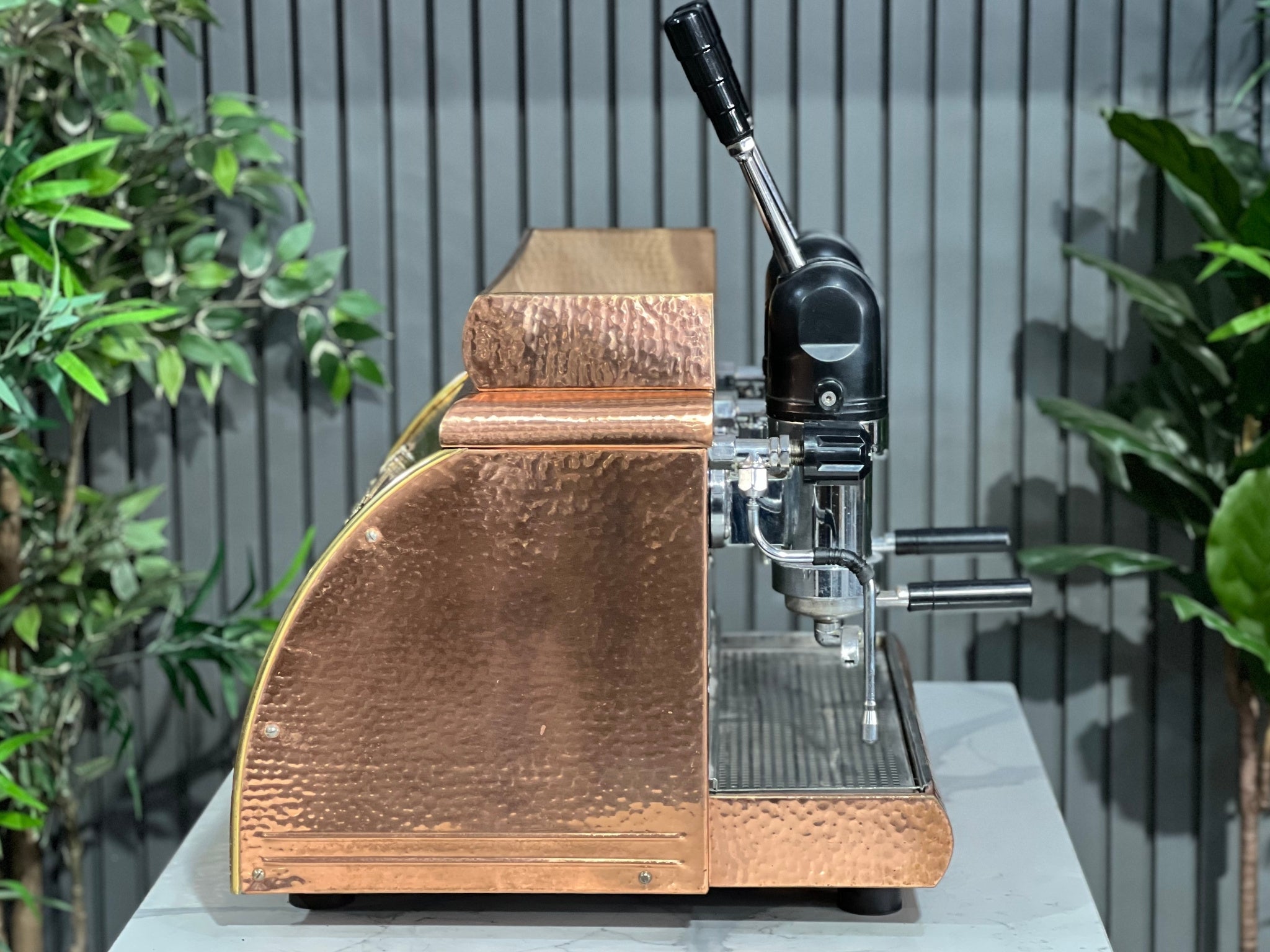 Victoria Arduino Athena Leva 2 Group Coffee Machine Bronze – Used espresso coffee machine side profile highlighting the lever mechanism, steam wand, and curvature of the bronze body against a marble bench backdrop.