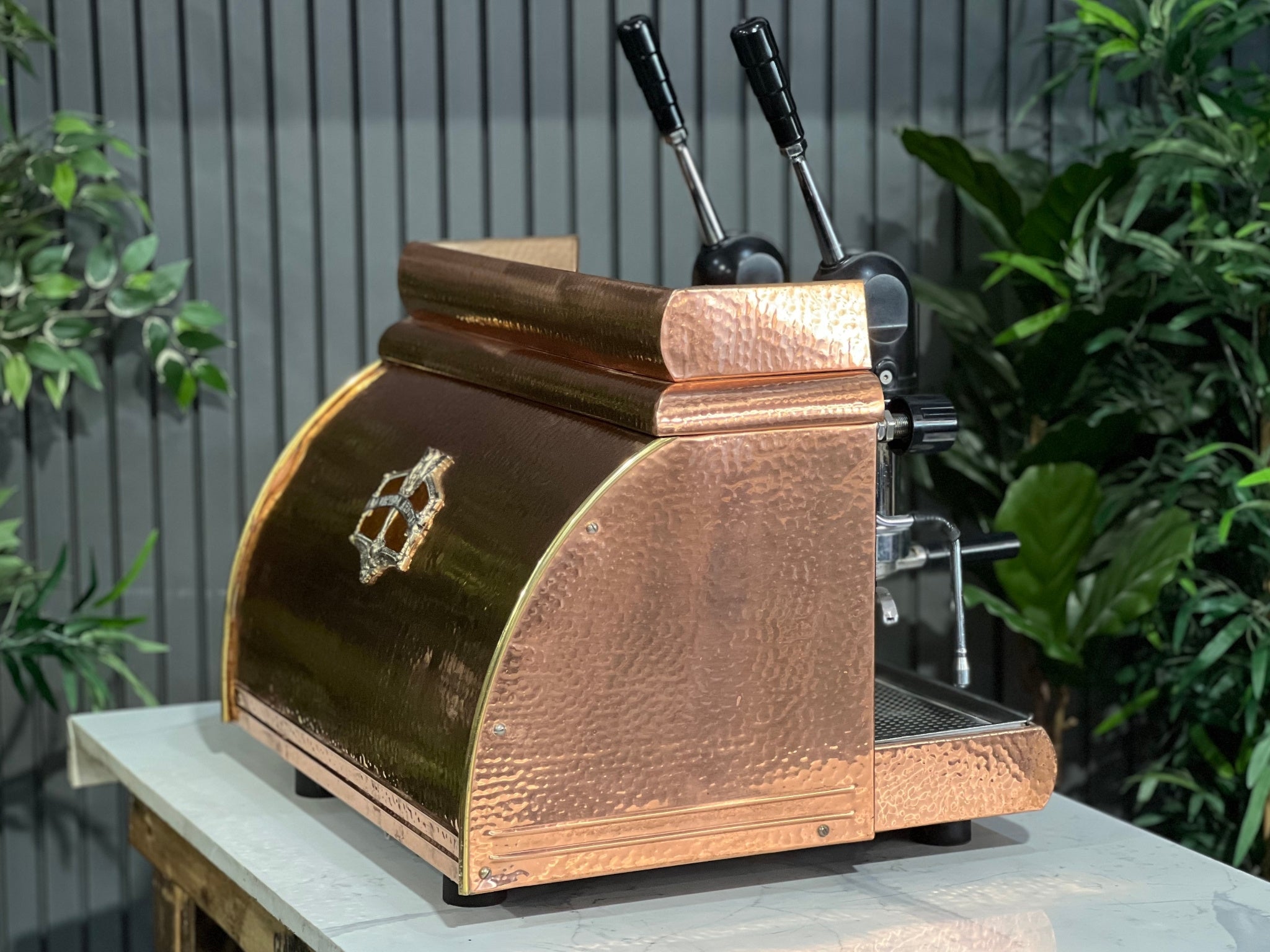 Victoria Arduino Athena Leva 2 Group Coffee Machine Bronze – Used espresso coffee machine shown at an angle on a marble bench with greenery in the background, highlighting its bronze body, dual group heads, lever arms, steam wands, and hot water tap.