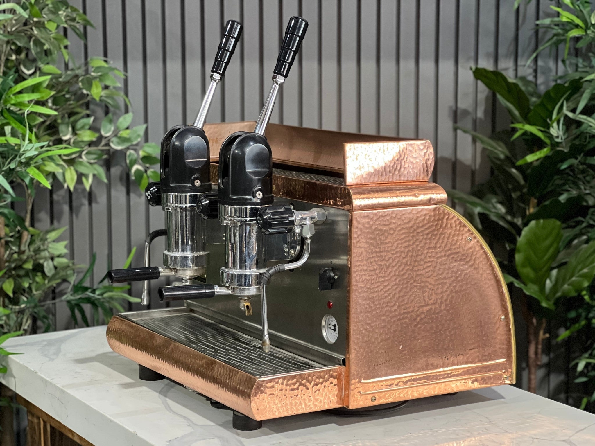 Victoria Arduino Athena Leva 2 Group Coffee Machine Bronze – Used espresso coffee machine shown at an angle on a marble bench with greenery in the background, highlighting its bronze body, dual group heads, lever arms, steam wands, and hot water tap.