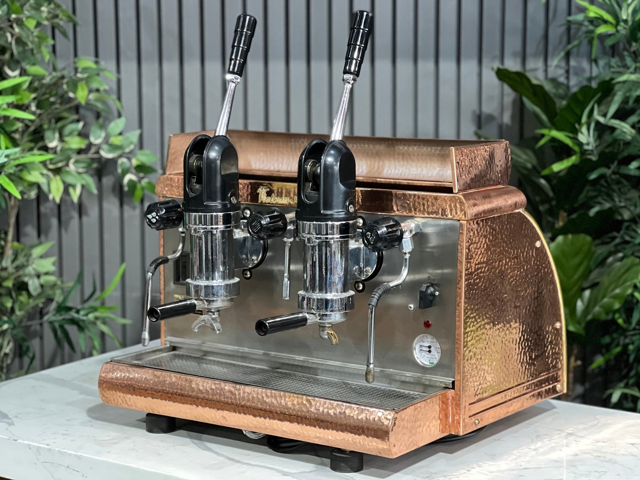Victoria Arduino Athena Leva 2 Group Coffee Machine Bronze – Used espresso coffee machine shown at an angle on a marble bench with greenery in the background, highlighting its bronze body, dual group heads, lever arms, steam wands, and hot water tap.
