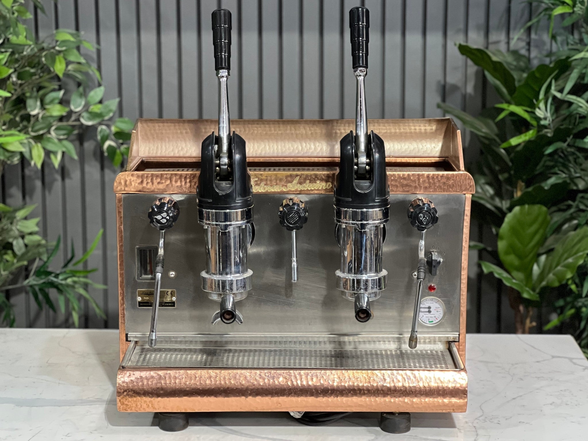 Victoria Arduino Athena Leva 2 Group Coffee Machine Bronze – Used espresso coffee machine shown at an angle on a marble bench with greenery in the background, highlighting its bronze body, dual group heads, lever arms, steam wands, and hot water tap.