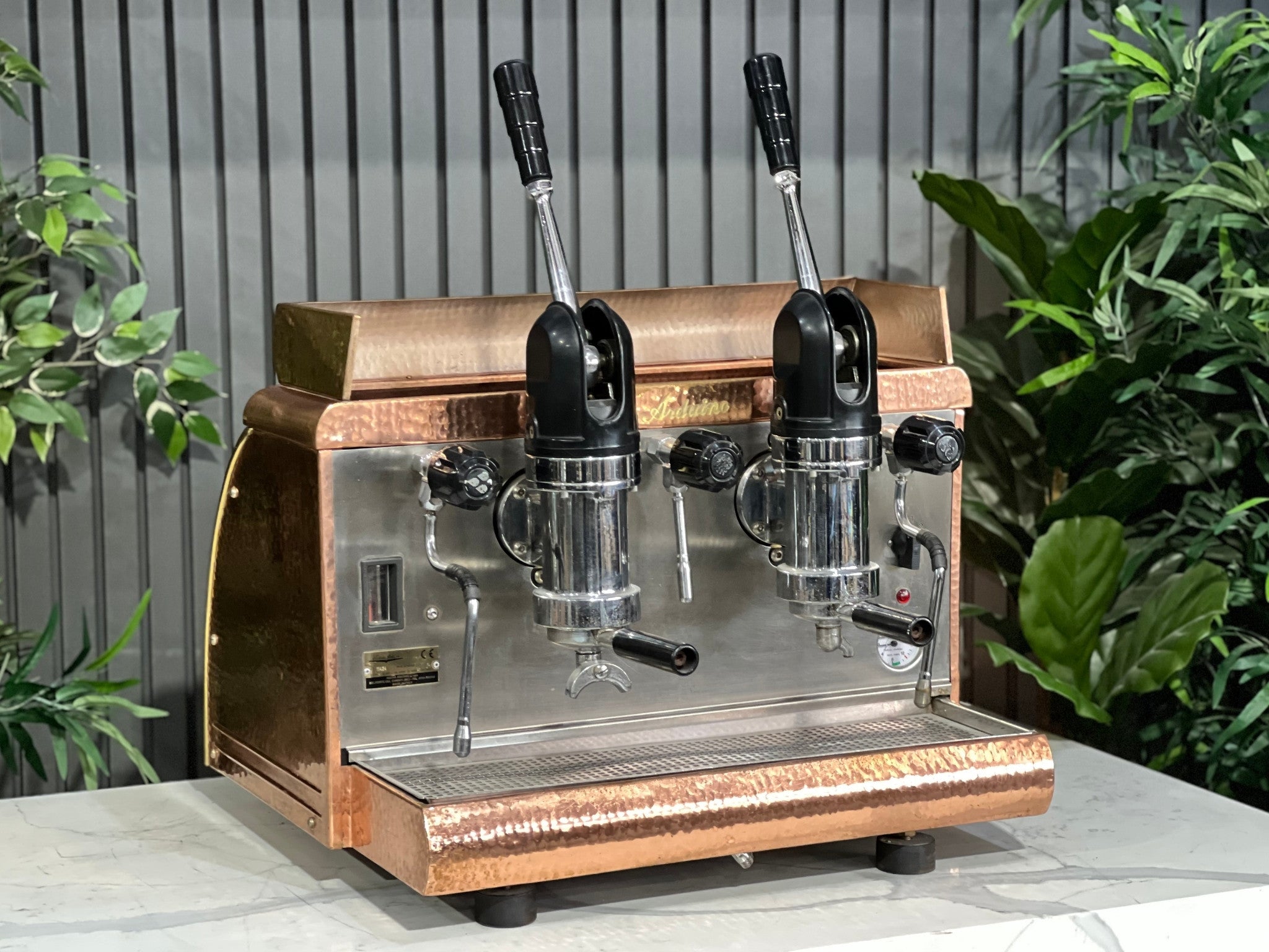 Victoria Arduino Athena Leva 2 Group Coffee Machine Bronze – Used espresso coffee machine shown at an angle on a marble bench with greenery in the background, highlighting its bronze body, dual group heads, lever arms, steam wands, and hot water tap.