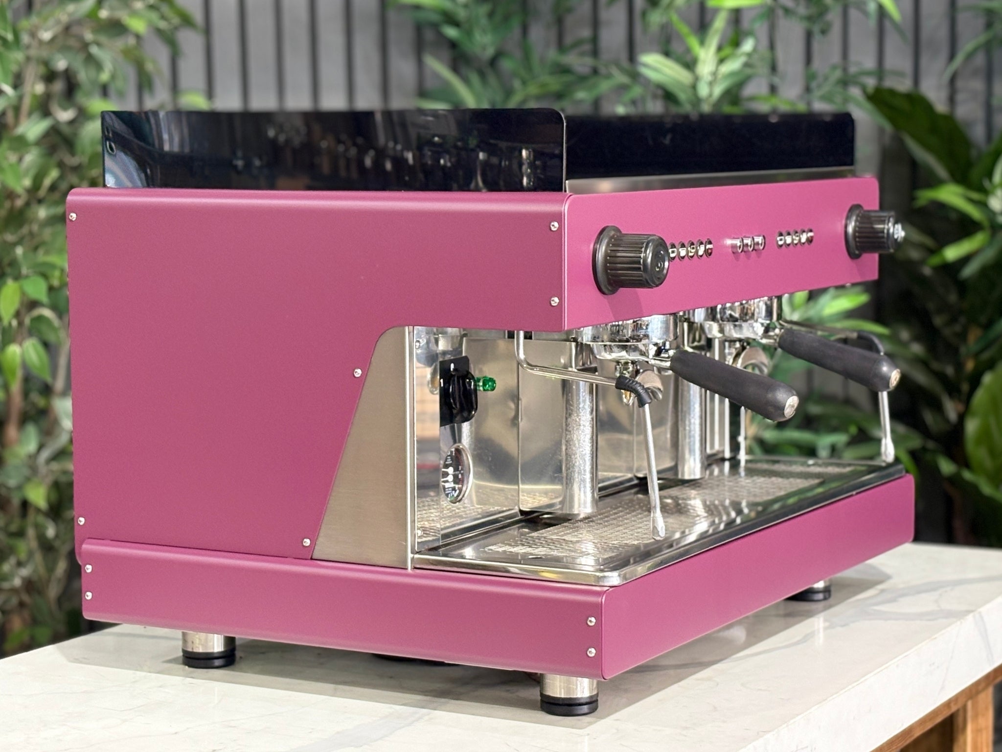 Wega Pegaso EVD 2 Group Coffee Machine Deep Pink – Used espresso coffee machine angled view showing E61 group heads, dual steam wands, and cup tray, deep pink stainless body, black acrylic top shroud, and stainless steel back panel, on a marble bench with greenery.