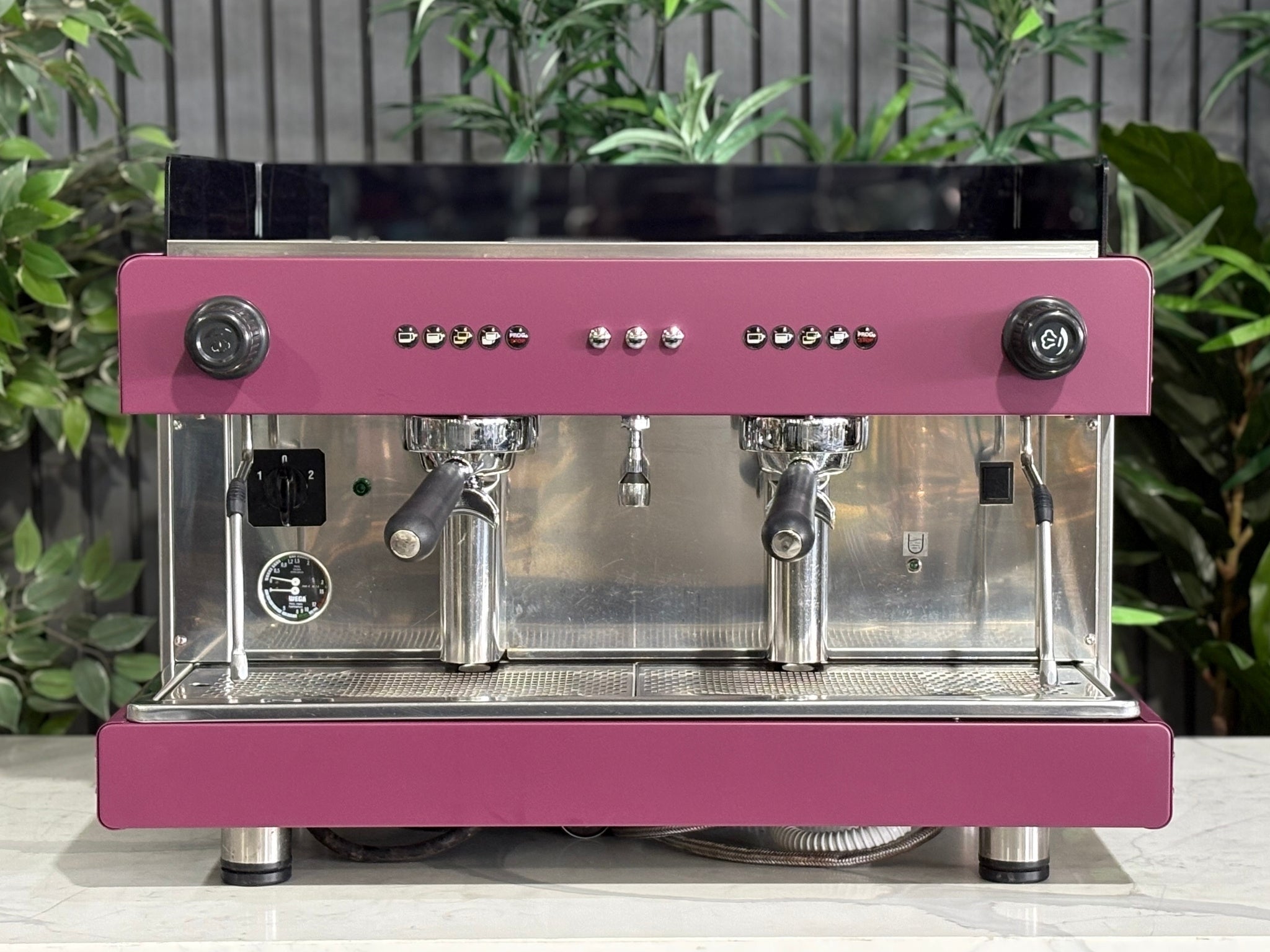 Wega Pegaso EVD 2 Group Coffee Machine Deep Pink – Used espresso coffee machine front view displaying control panel, twin group heads, steam wands, cup tray, deep pink stainless body, black acrylic top shroud, on a marble bench with plants behind.