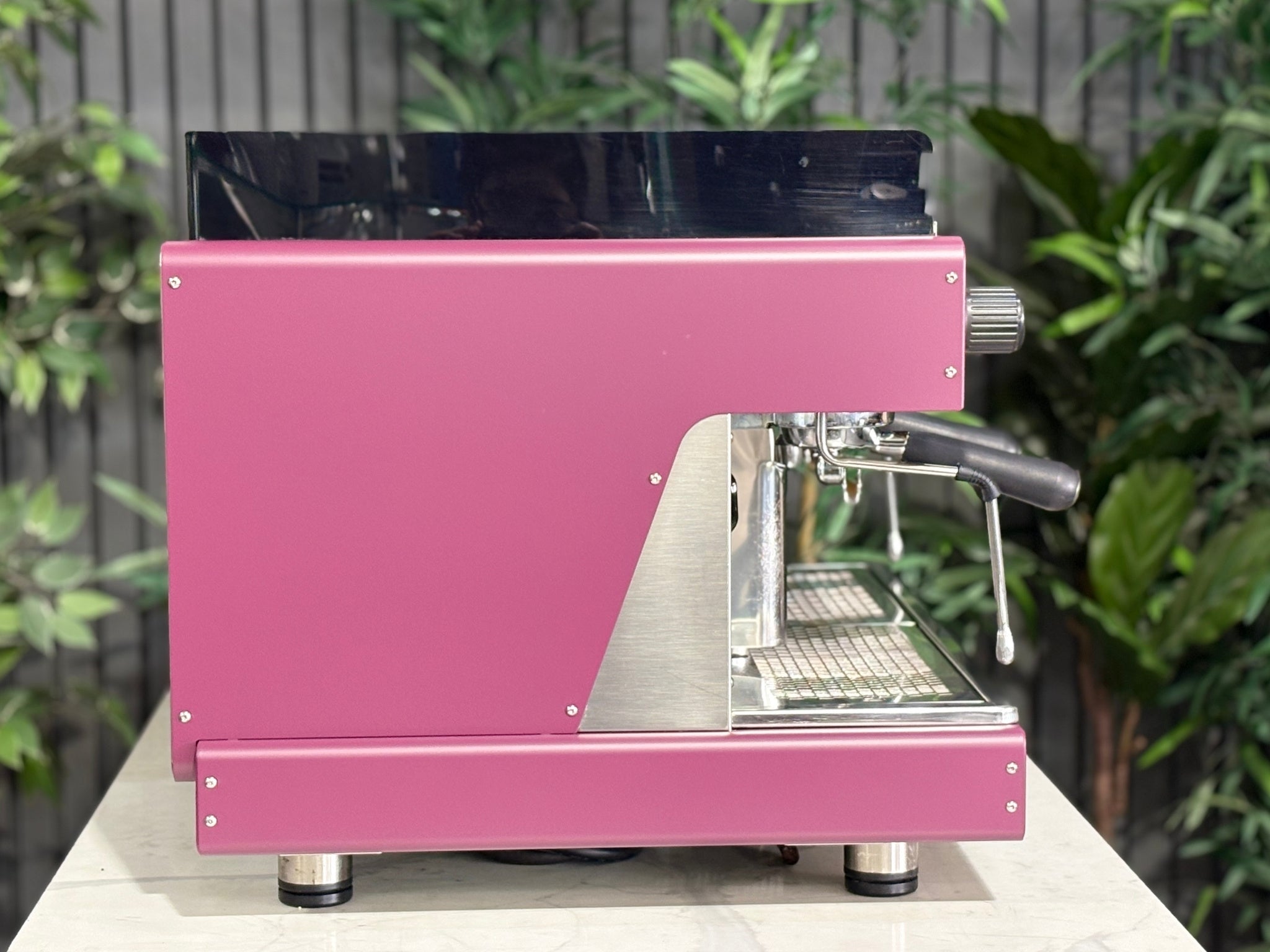 Wega Pegaso EVD 2 Group Coffee Machine Deep Pink – Used espresso coffee machine side view showing flat deep pink stainless panel, black acrylic top shroud, and black accents, on a marble bench with greenery behind.